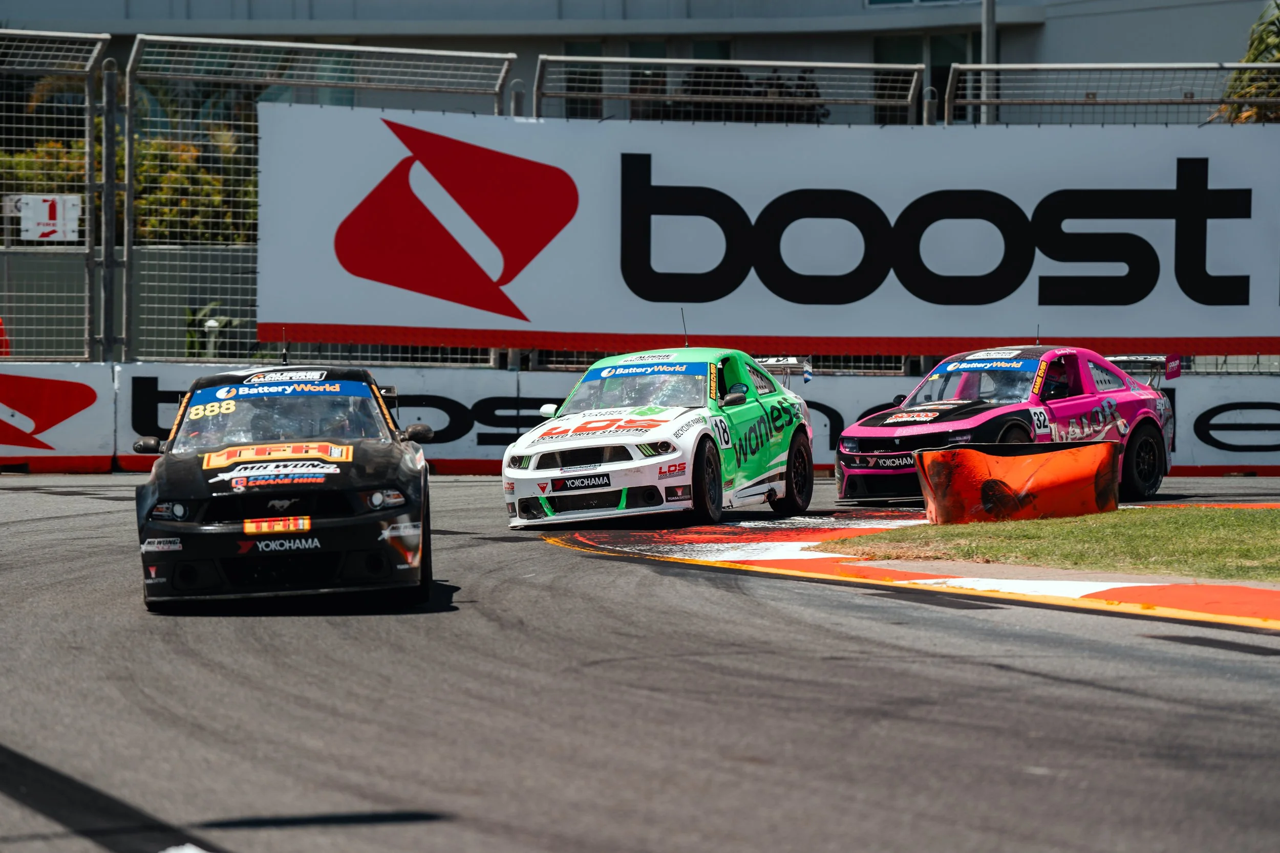 Three race cars on a track, with a large 'boost' sign in the background. The cars are navigating a turn, with the middle car partly off the track and close to grass. The car on the left is black with orange and white branding, the middle car is green