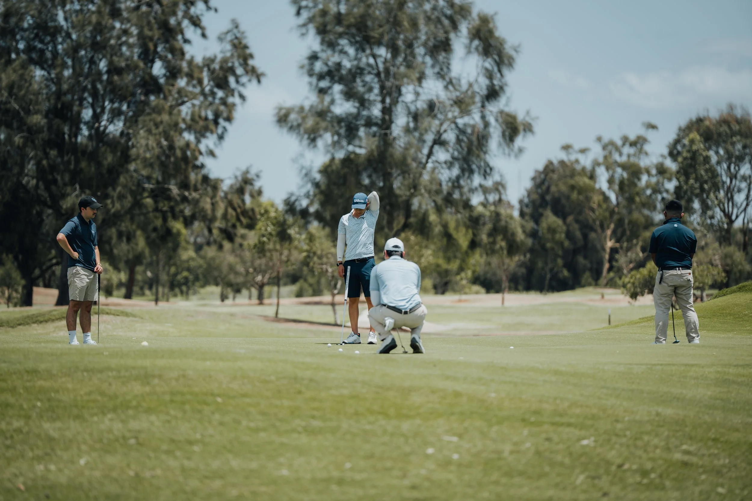 Group of four men on a golf course, with two preparing to putt and others observing, surrounded by trees under a partly cloudy sky.