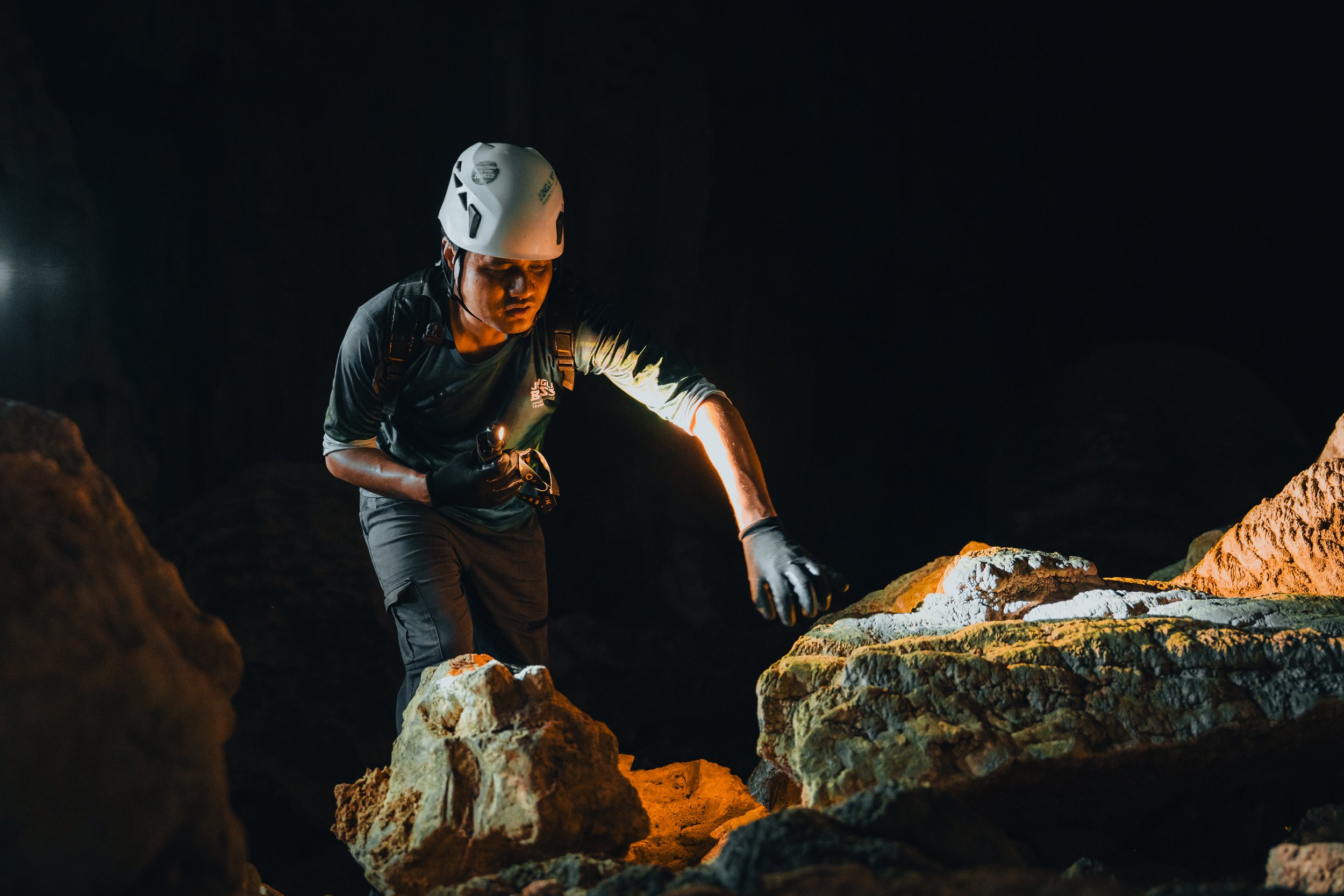 A person wearing a helmet and gloves exploring a dark cave with rocks, using a flashlight to illuminate the surroundings.
