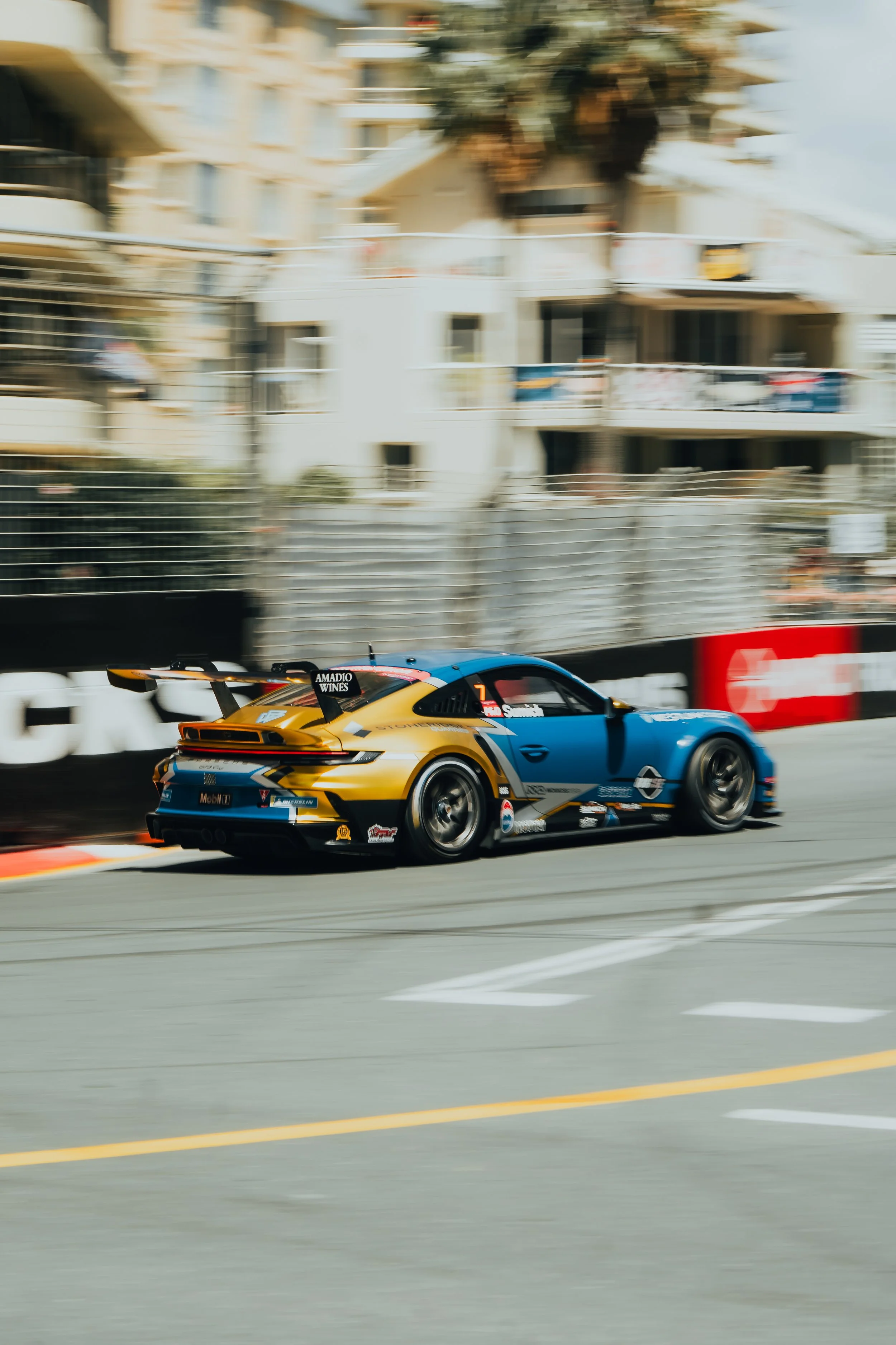A blue and yellow race car speeding on a track with blurred background of buildings and trees.