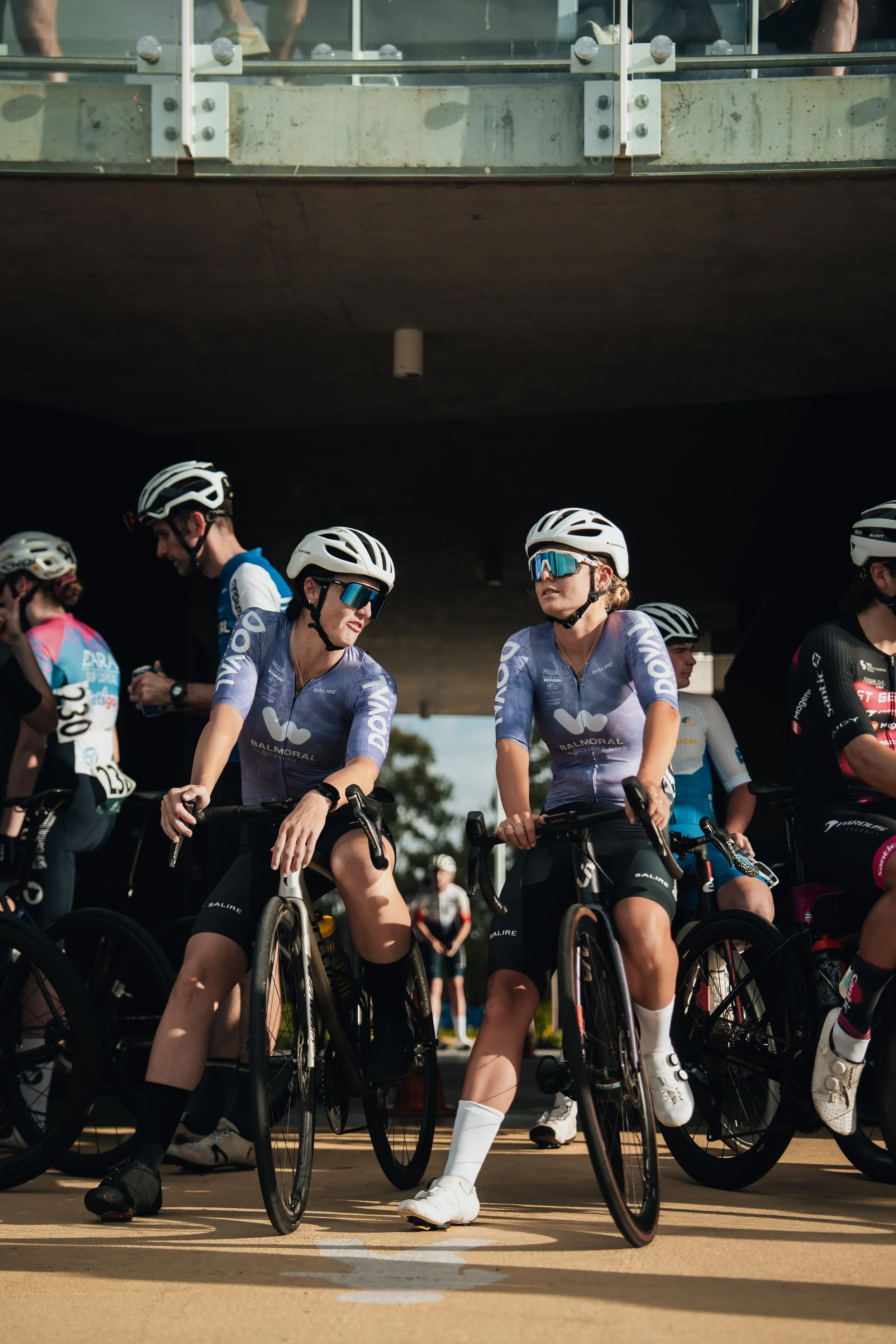 Cyclists in team uniforms and helmets preparing to race at the starting line under a bridge.
