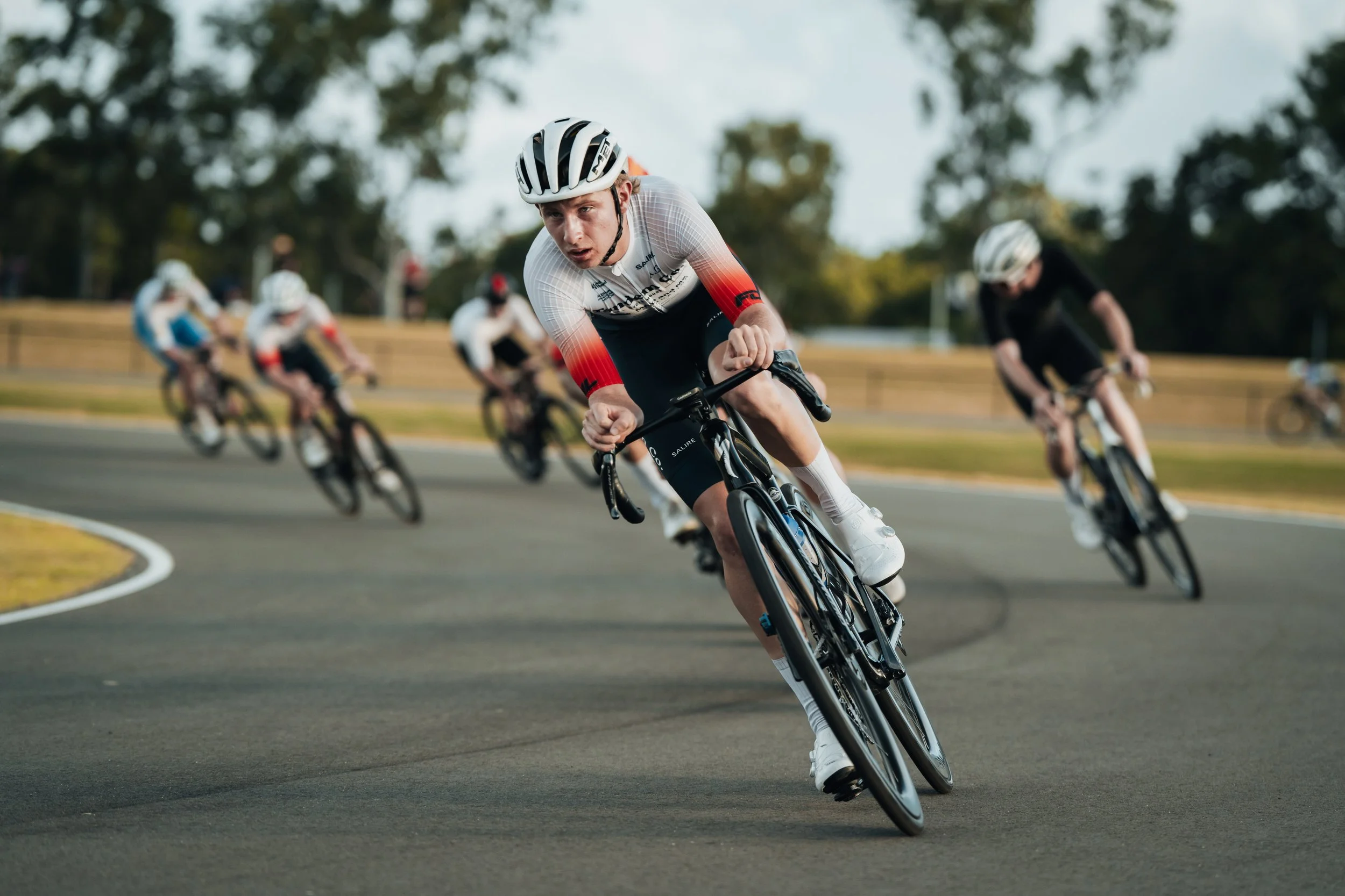 Cyclists racing on a paved outdoor track, leaning into a curve with trees in the background.