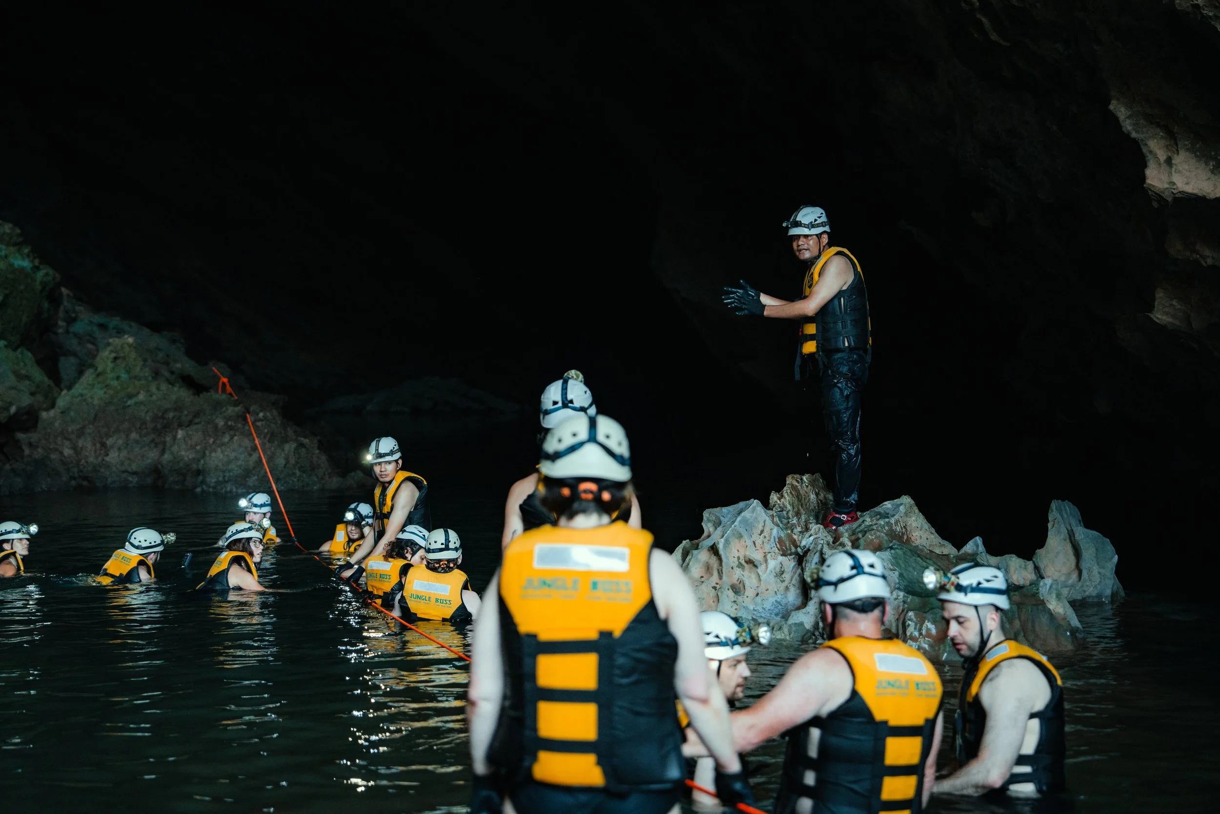 Group of people in life jackets and helmets participating in a caving or spelunking activity inside a dark cave, with one person standing on a rock and explaining to the others.