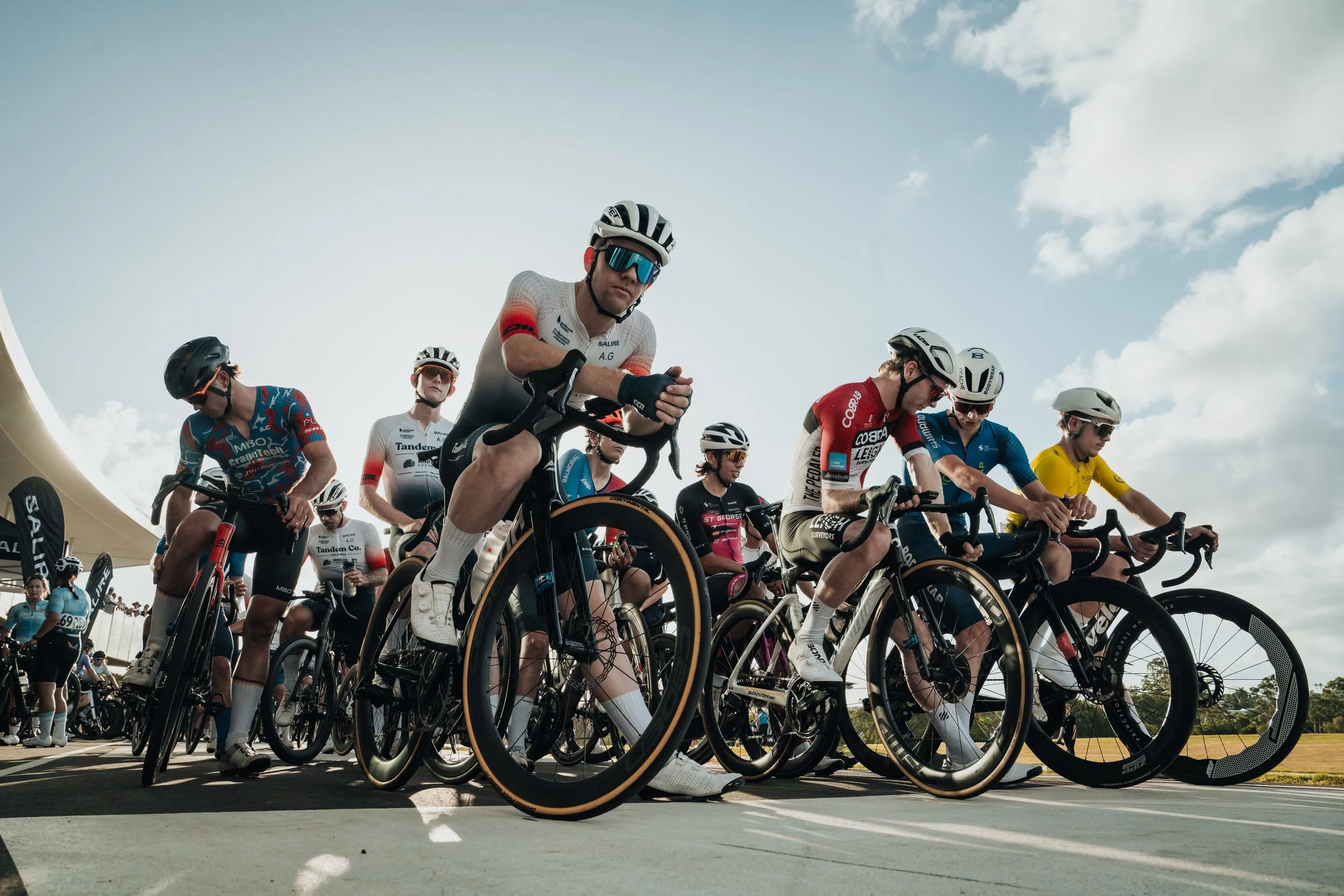Group of cyclists at the starting line of a race, wearing helmets and racing gear, ready to begin.