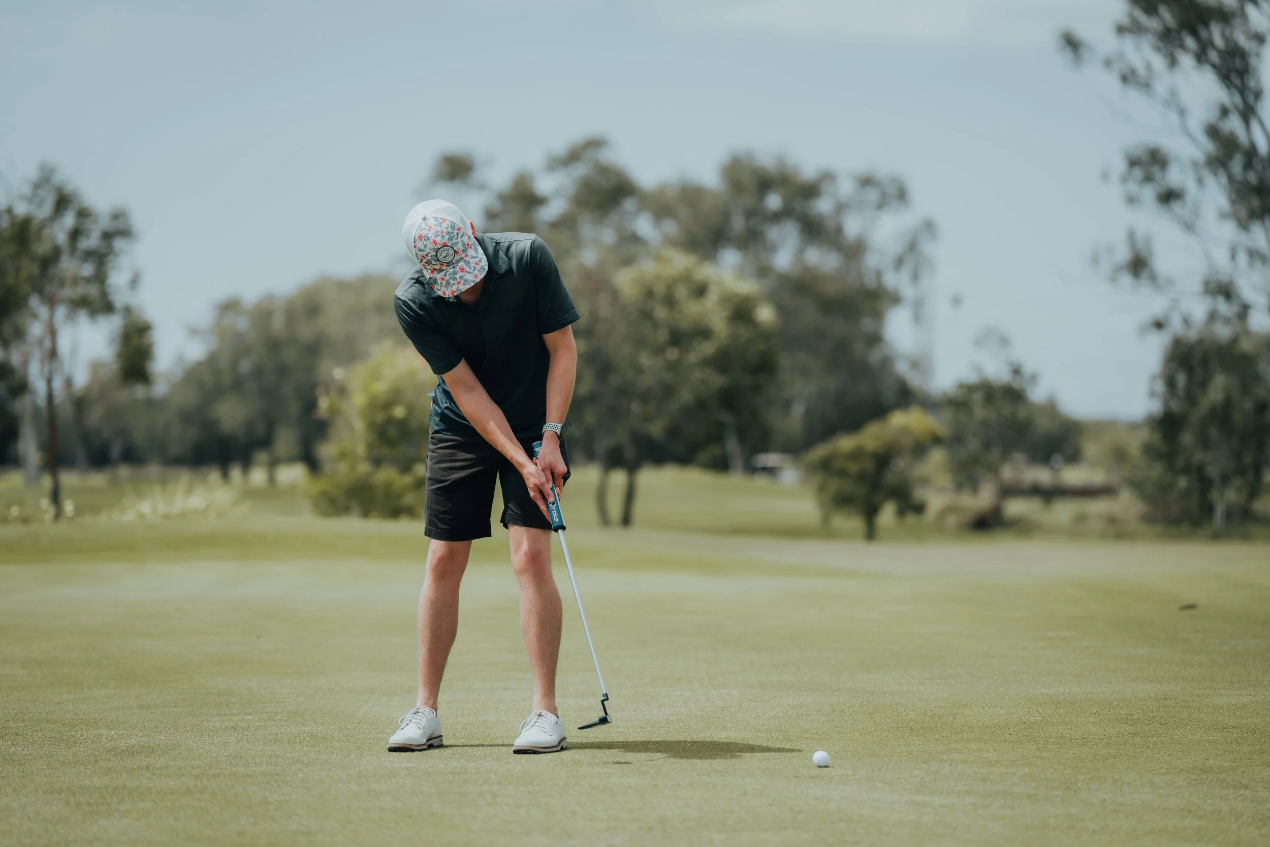 Man wearing a hat, black shirt, black shorts, and white shoes putting on a golf course