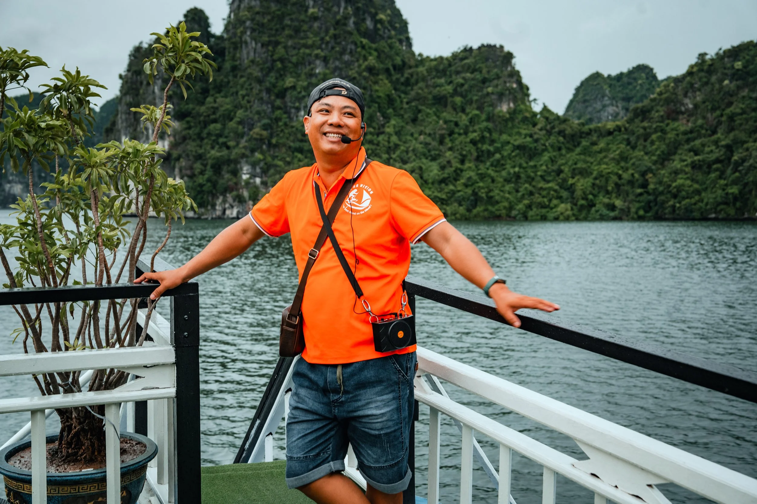 A smiling man in an orange shirt with a headset, standing on a boat deck with a lake and green mountains in the background.