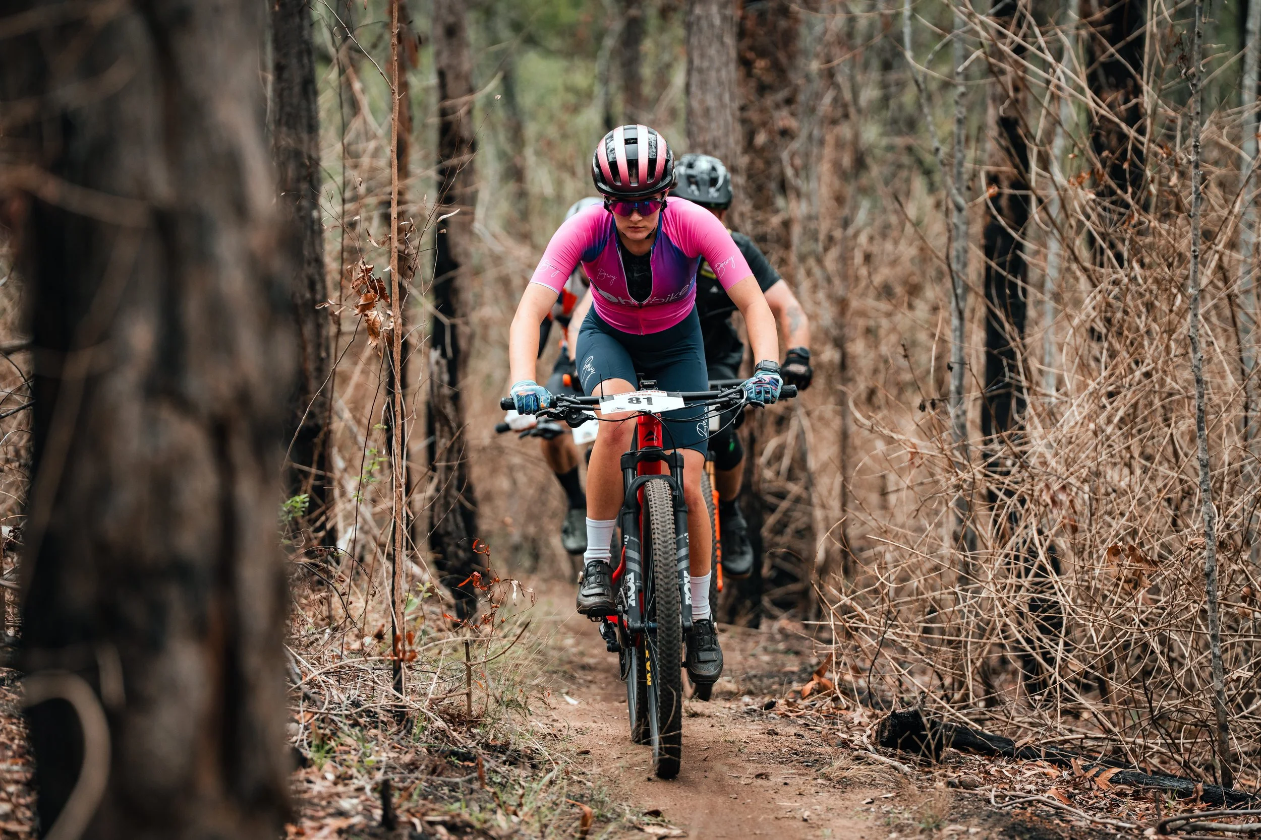A woman riding a mountain bike through a narrow dirt trail in a wooded area, wearing a pink and purple jersey, helmet, and sunglasses, with two other cyclists in the background.