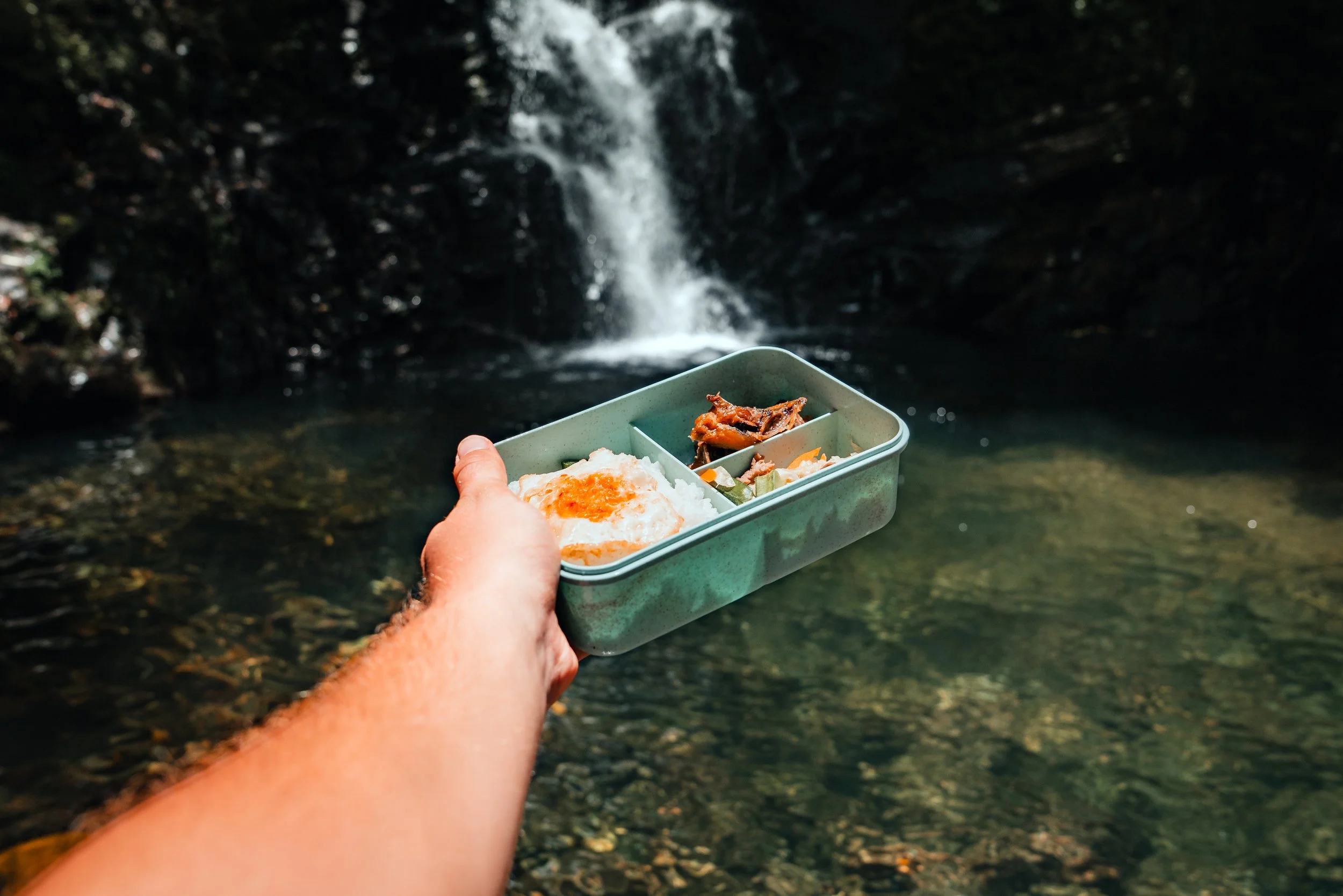 Person holding a green lunchbox with food, including rice with fried egg, near a small waterfall in a natural setting.