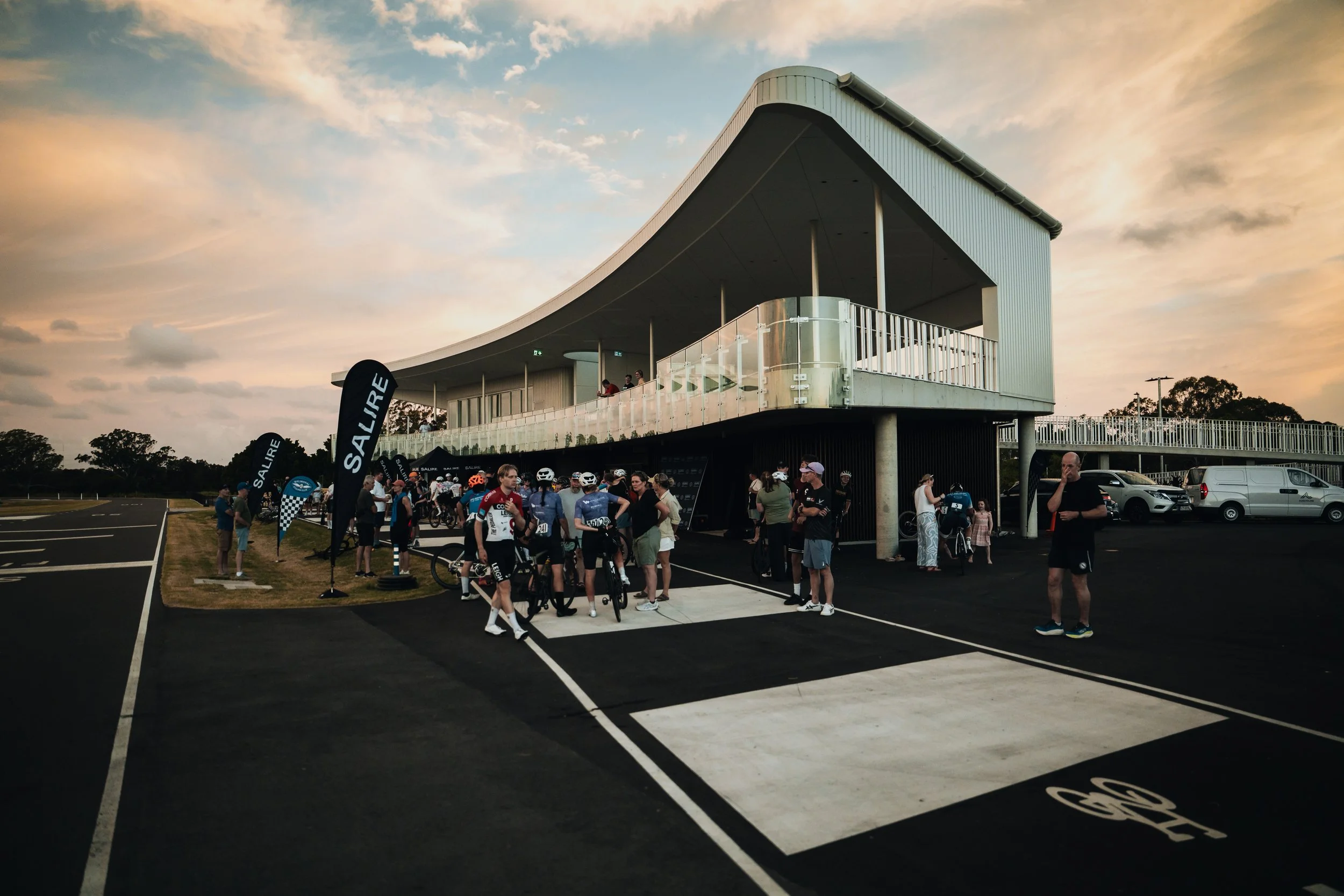 People gathering outside a modern building adjacent to a parking lot, possibly participating in a cycling event, with flags displaying the word 'SAILRE' and a sunset sky in the background.
