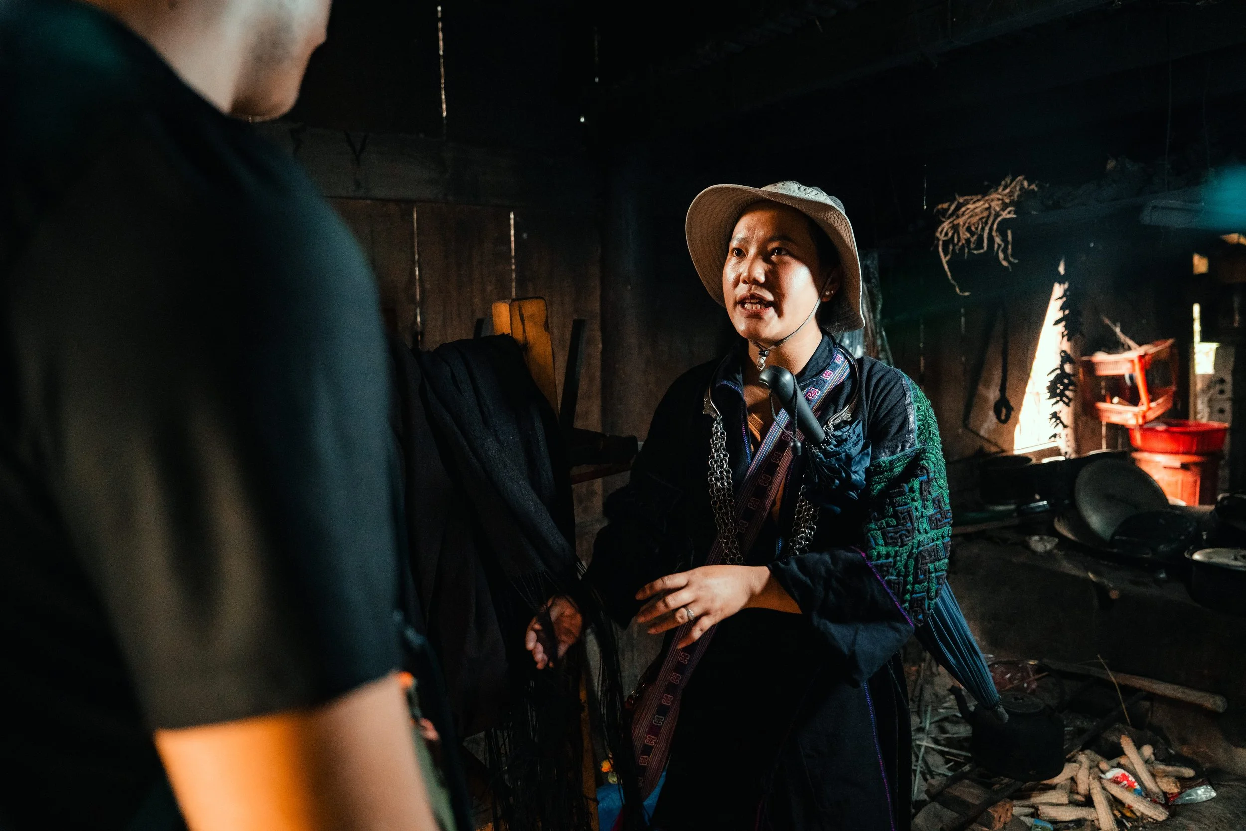A woman wearing a beige sun hat and dark traditional clothing speaking to another person inside a rustic, dimly lit room, with kitchen utensils and red containers visible in the background.