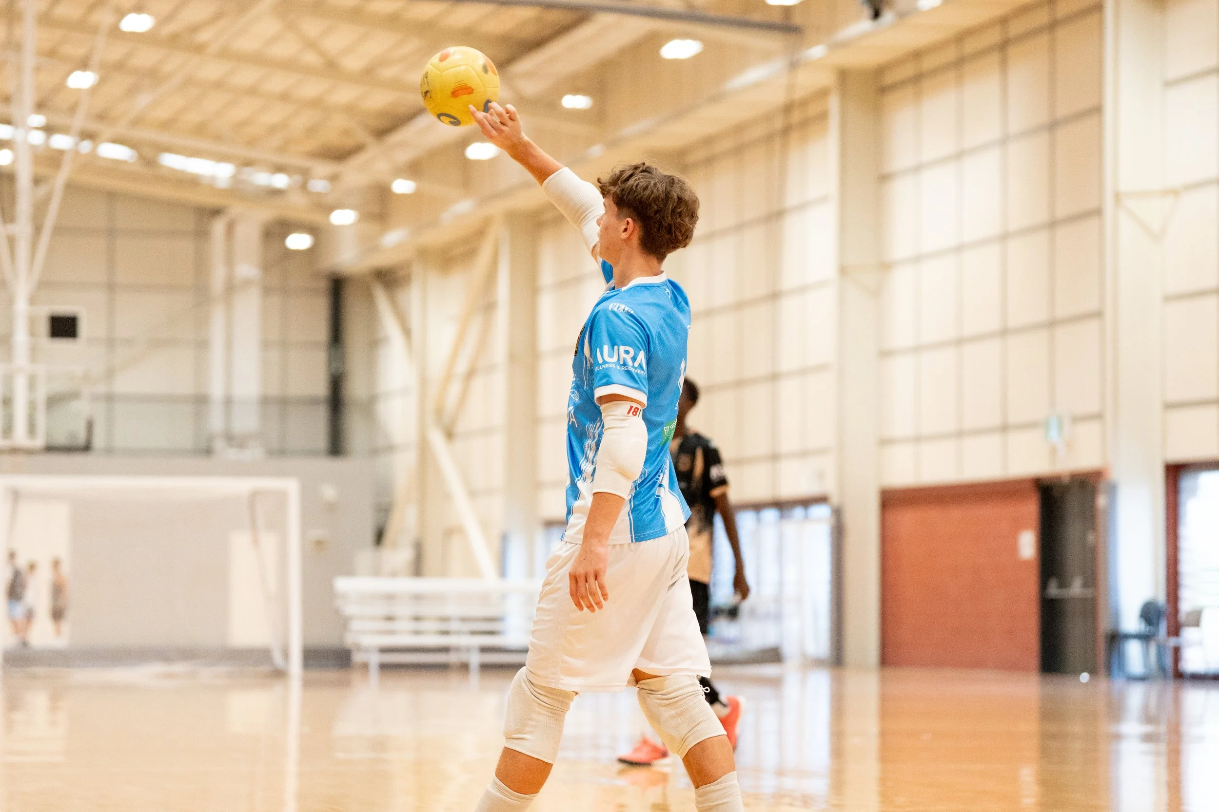 A young man in a blue sports jersey and white shorts playing indoor volleyball, preparing to serve or spike the ball inside a gymnasium with a wooden floor and large windows.