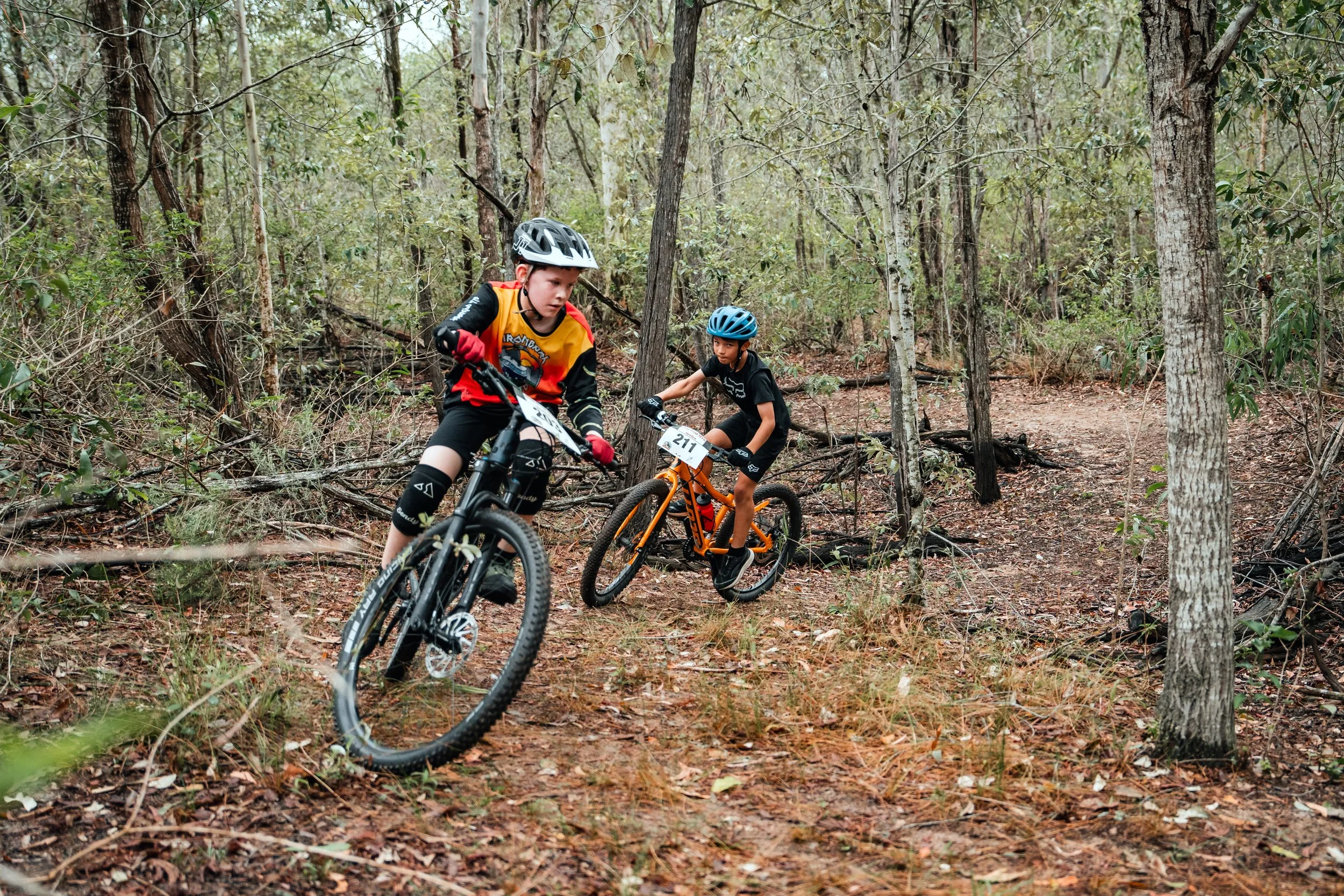 Two boys riding mountain bikes through a wooded trail with trees and dry leaves.