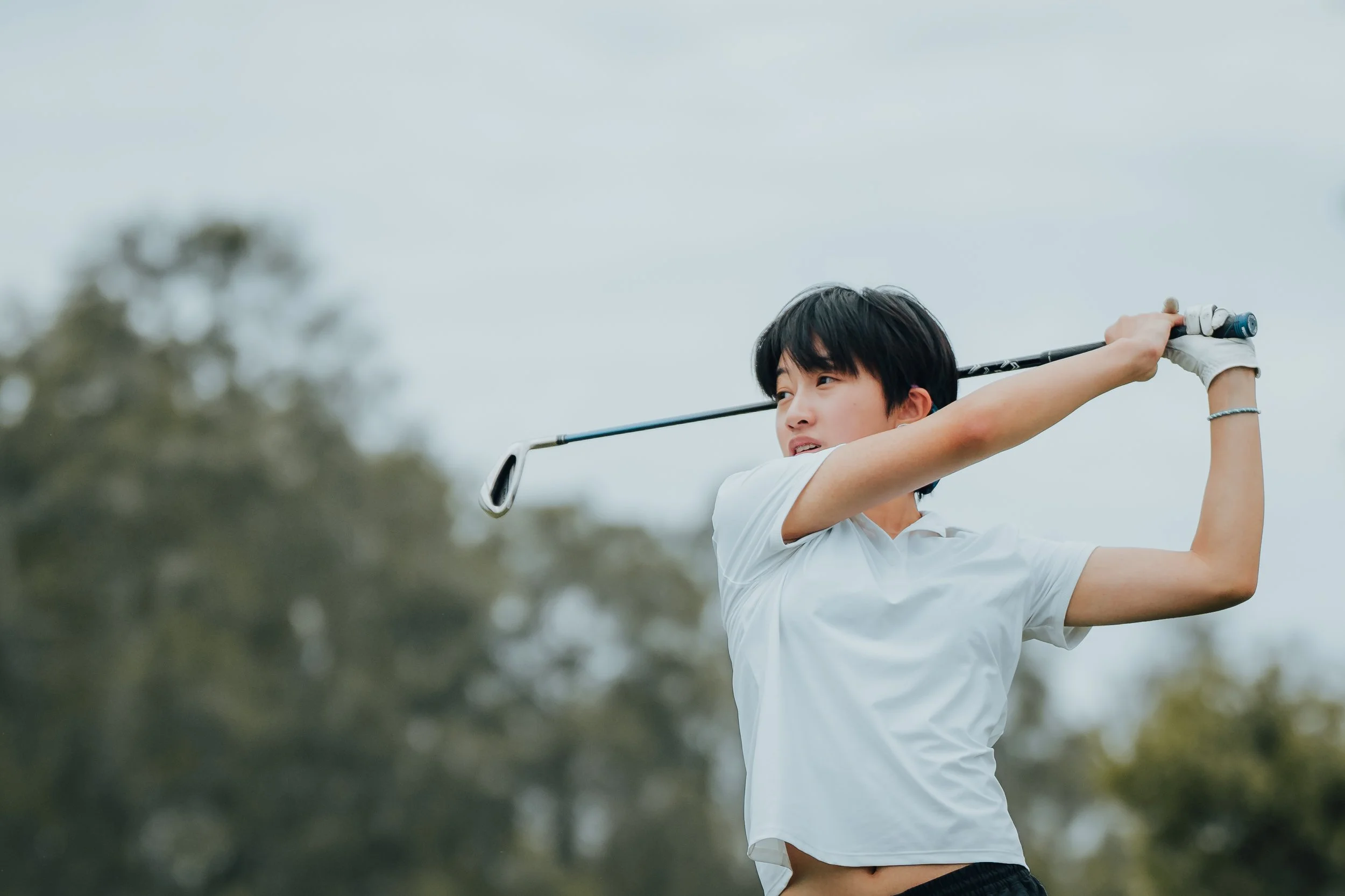 Young woman in white shirt and black shorts swinging a golf club on a golf course with trees in the background.