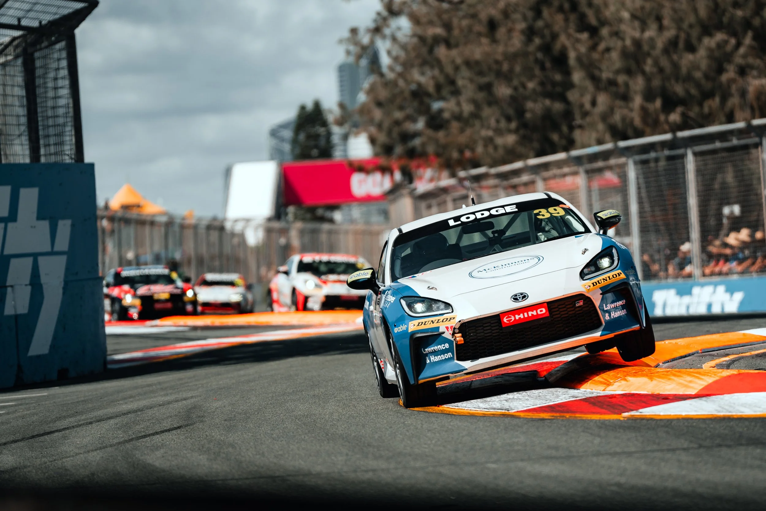 A race car is airborne while taking a sharp turn on a race track with other race cars following behind. The race cars are navigating a corner with orange and white curbs, and there are spectators watching from the stands. The sky is partly cloudy.