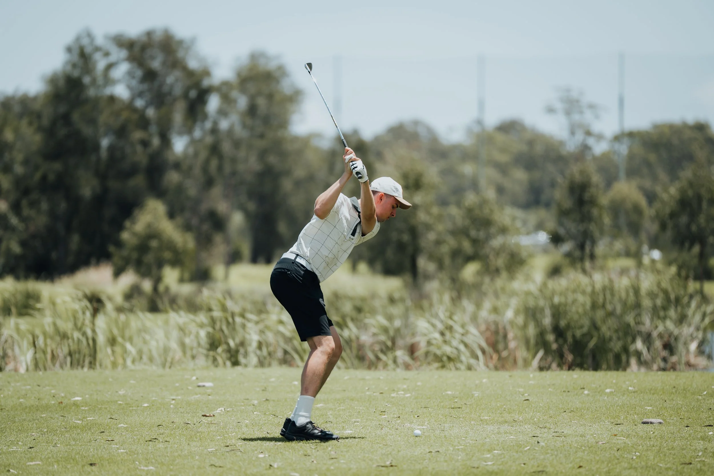 A man playing golf on a sunny day, preparing to swing his club, standing on a green golf course with trees in the background.