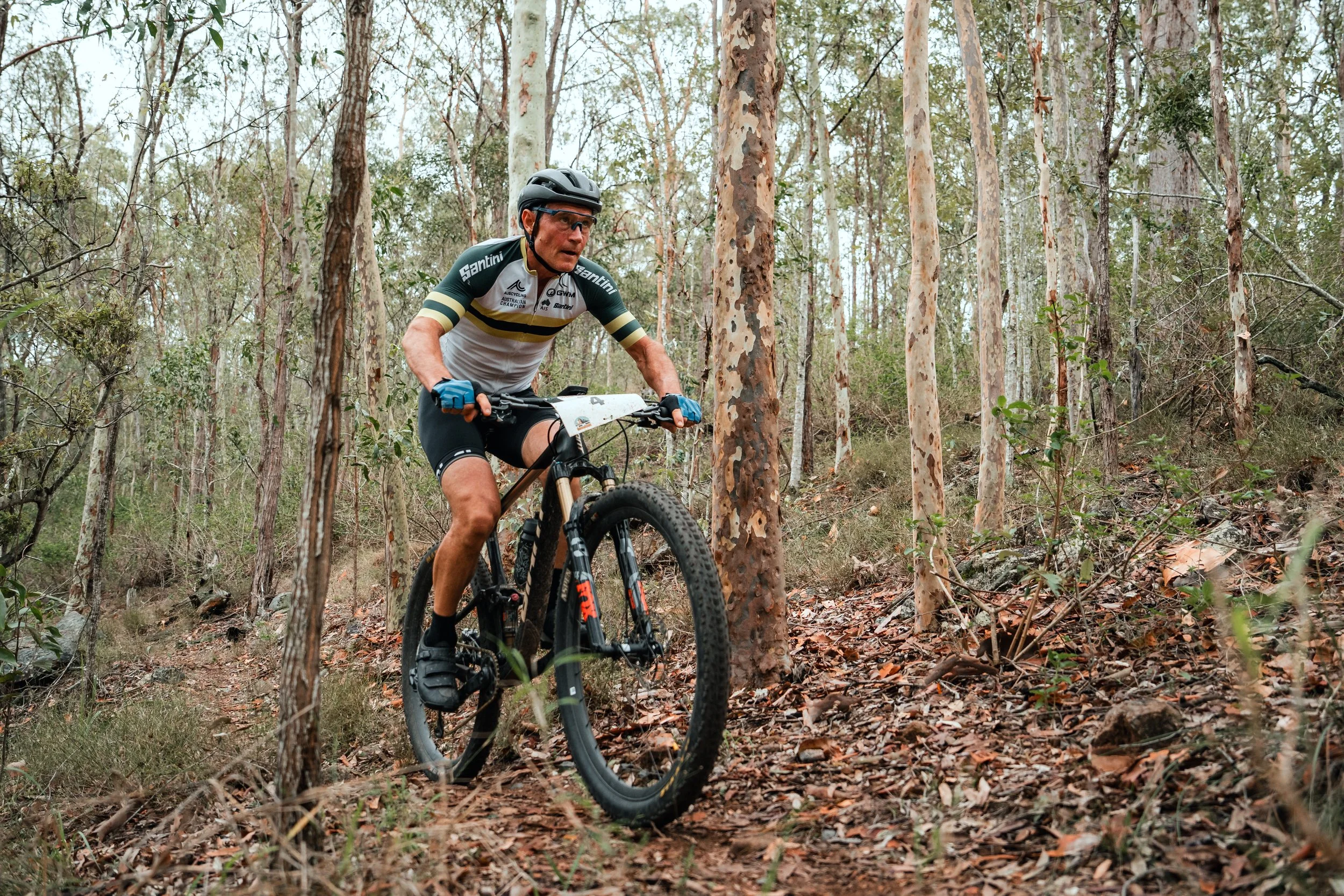 A man riding a mountain bike through a forest trail, wearing a helmet, cycling gloves, and a cycling jersey.
