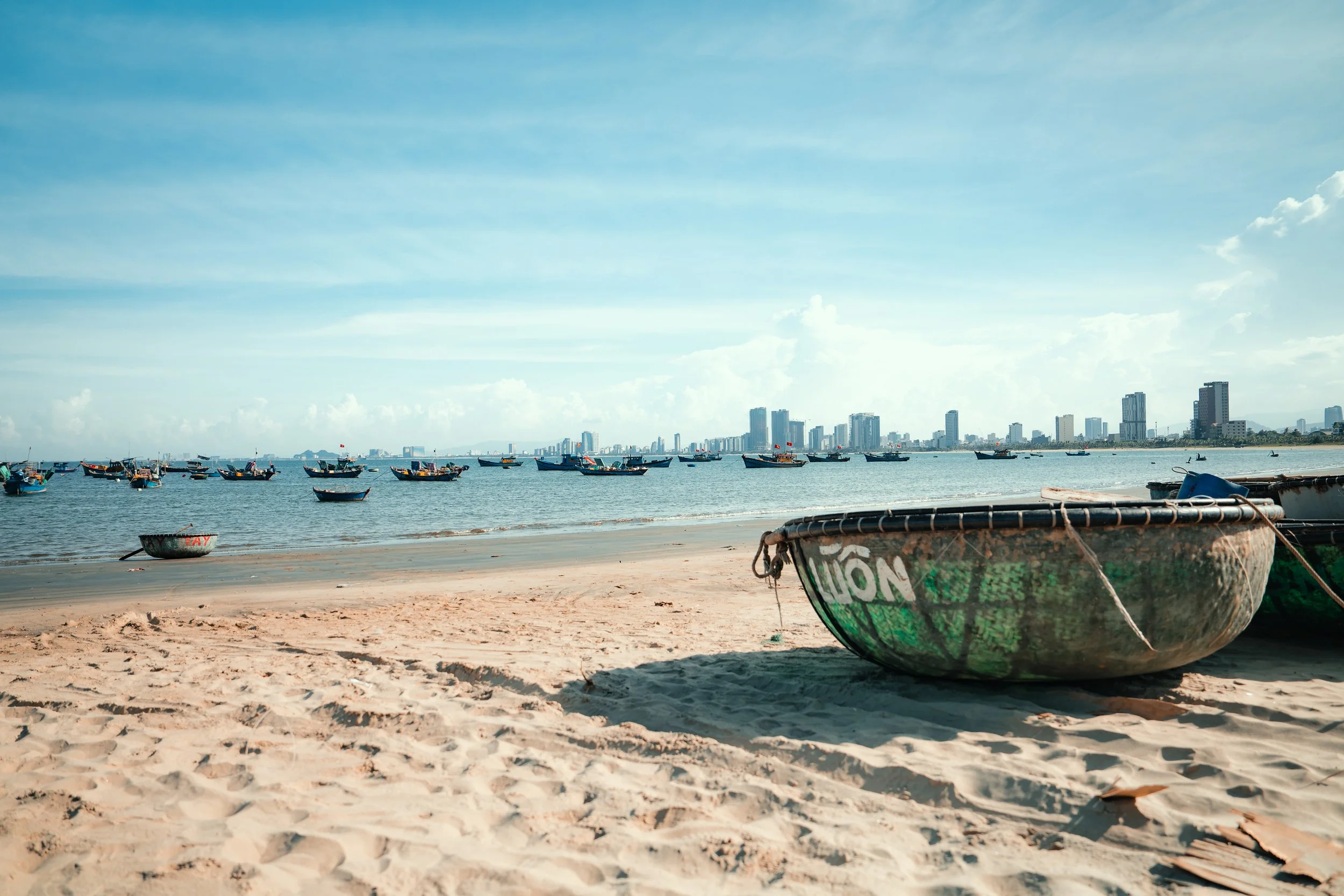 A beach scene with boats resting on the sandy shore, a city skyline with high-rise buildings in the background, and a clear blue sky with scattered clouds.