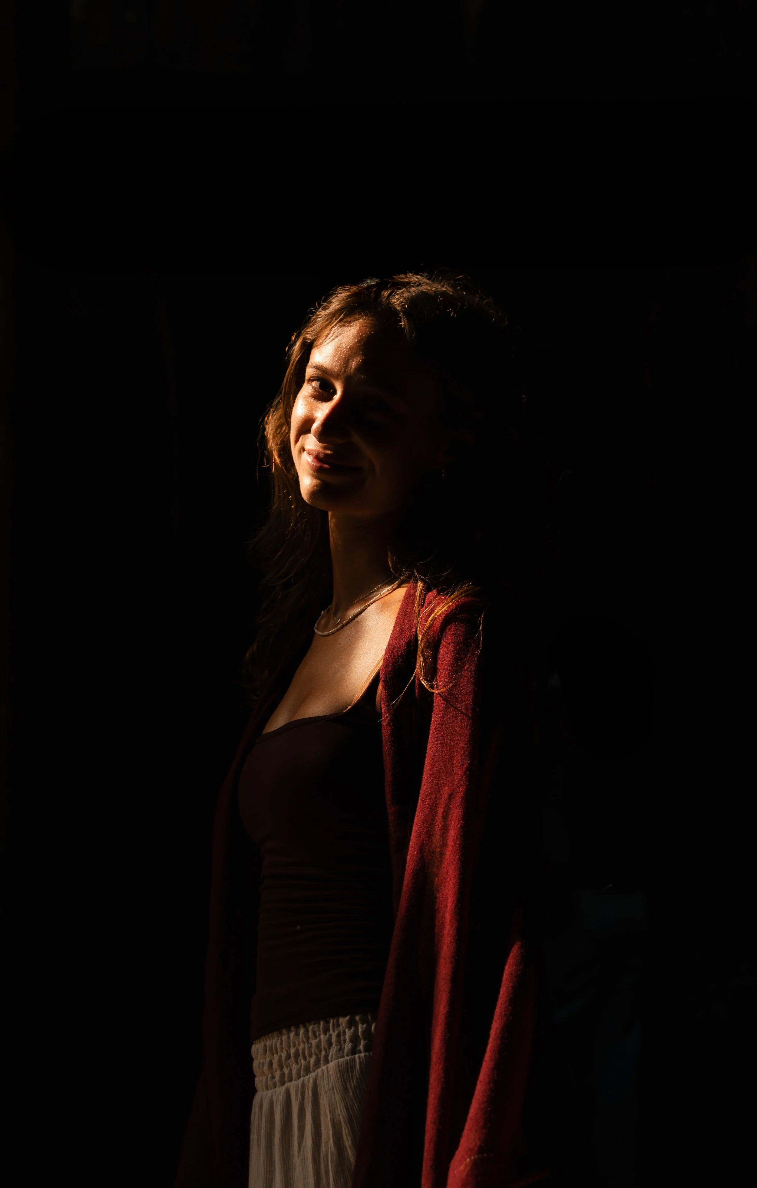 A woman with long wavy hair smiling, illuminated by a soft light from her right side, with a dark background.