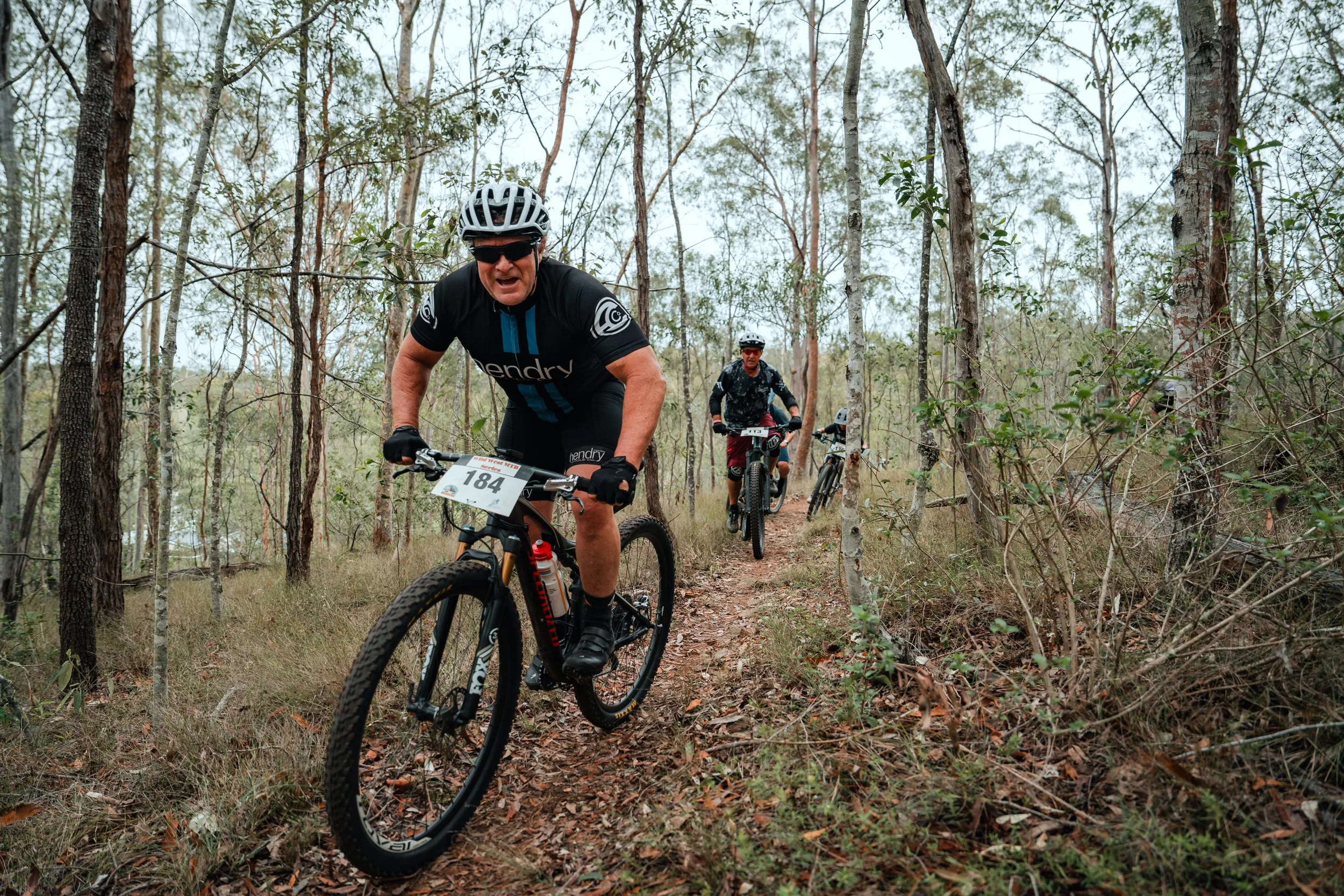 Three mountain bikers riding through a wooded trail, with the leading rider showing an intense expression, wearing a black helmet and cycling gear, with the other two following behind in the background.