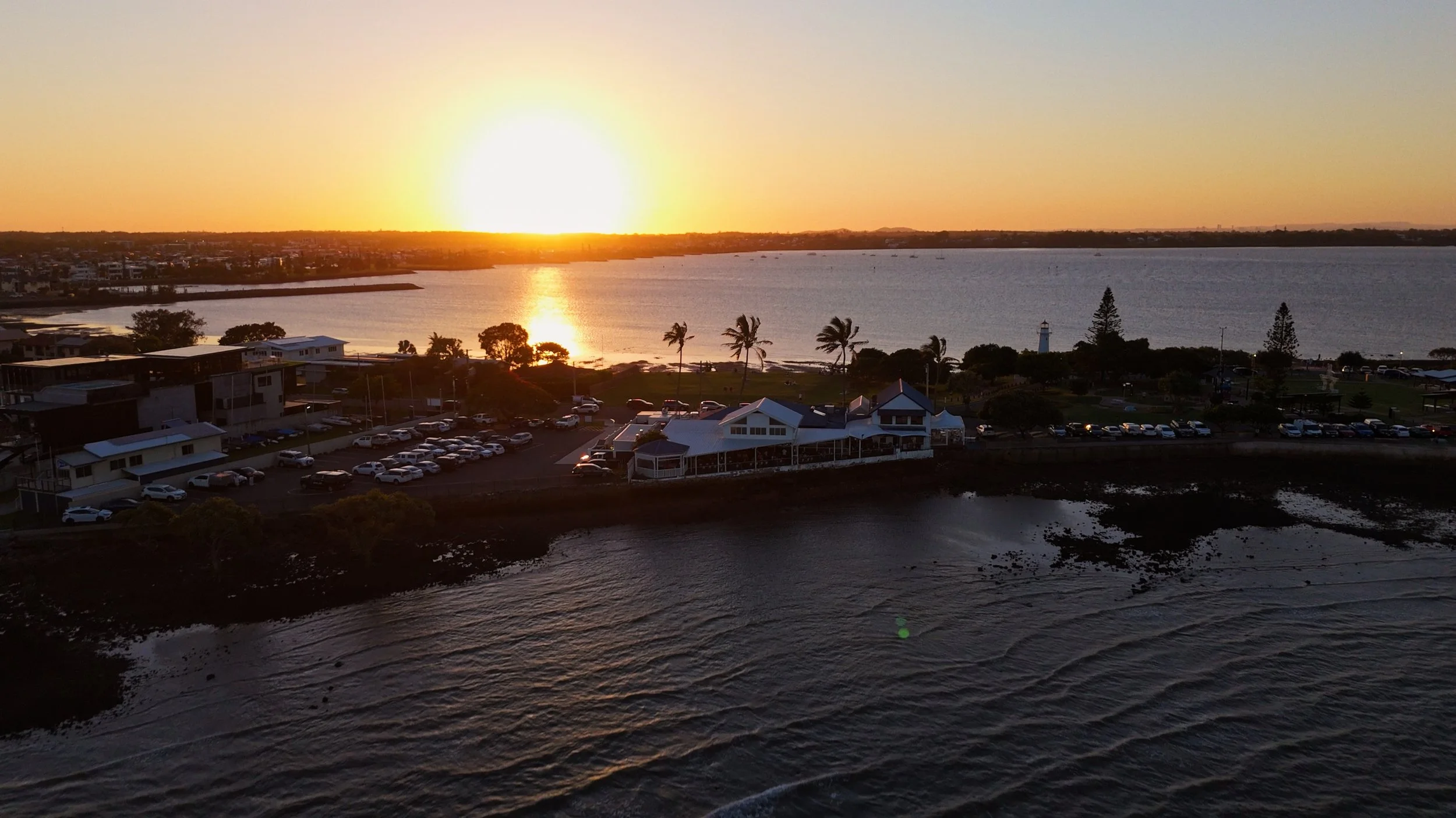 Sunset over a waterfront with palm trees, buildings, and a parking lot by the river.