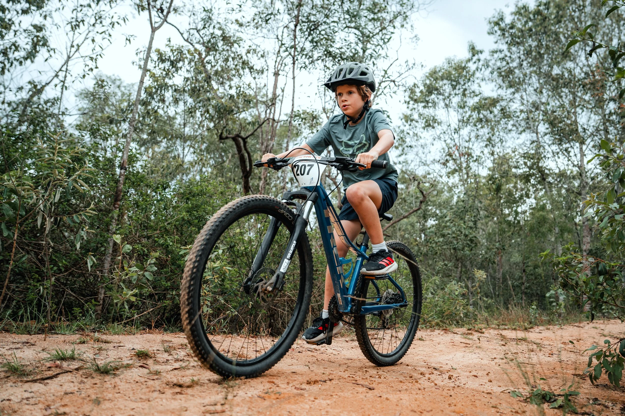 A boy riding a mountain bike on a dirt trail through a forest, wearing a helmet and sports gear.