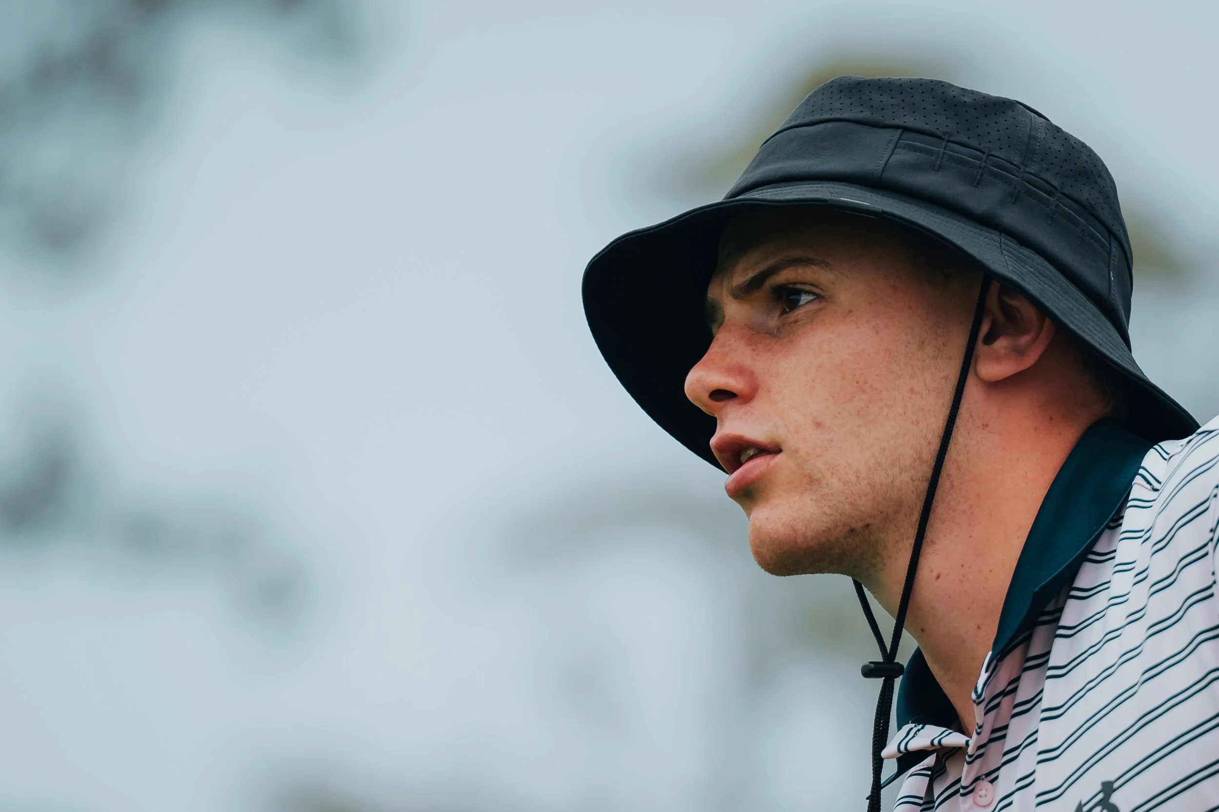 Close-up of a young man with fair skin, freckles, and short hair, wearing a black bucket hat and a striped shirt, looking intently to the right outdoors against a blurred sky background.