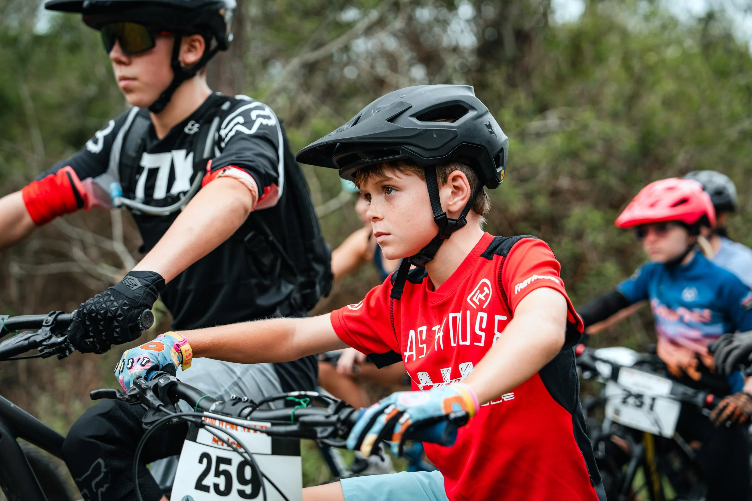 Young boys wearing helmets and riding mountain bikes on a trail in a forest.