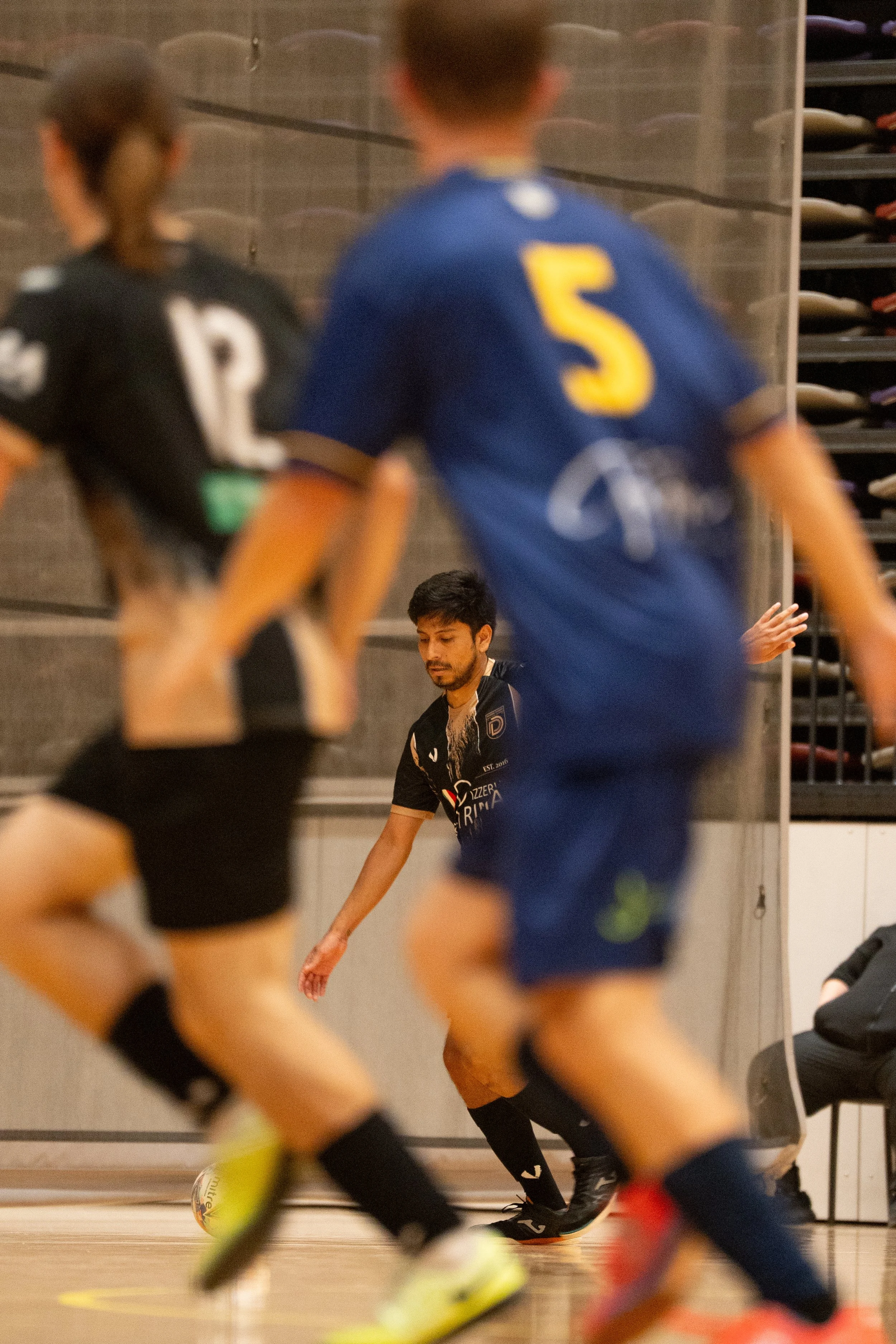 Indoor soccer game with two players in black and blue uniforms and a referee in black, with a man in black observing in the background.