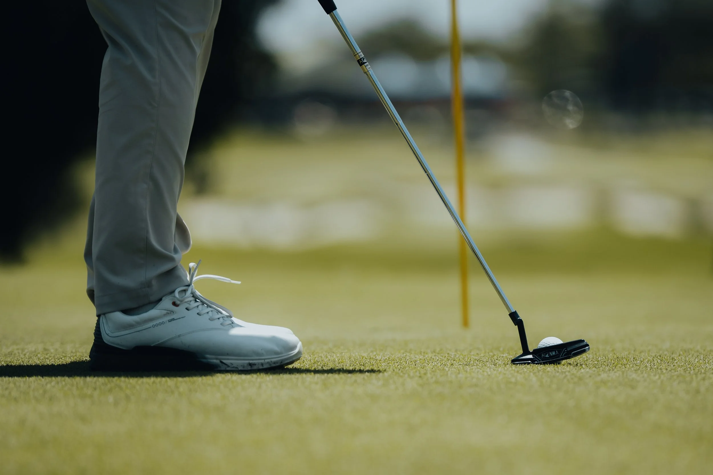 Close-up of a golfer's feet and golf club near a golf ball on the green putting surface.