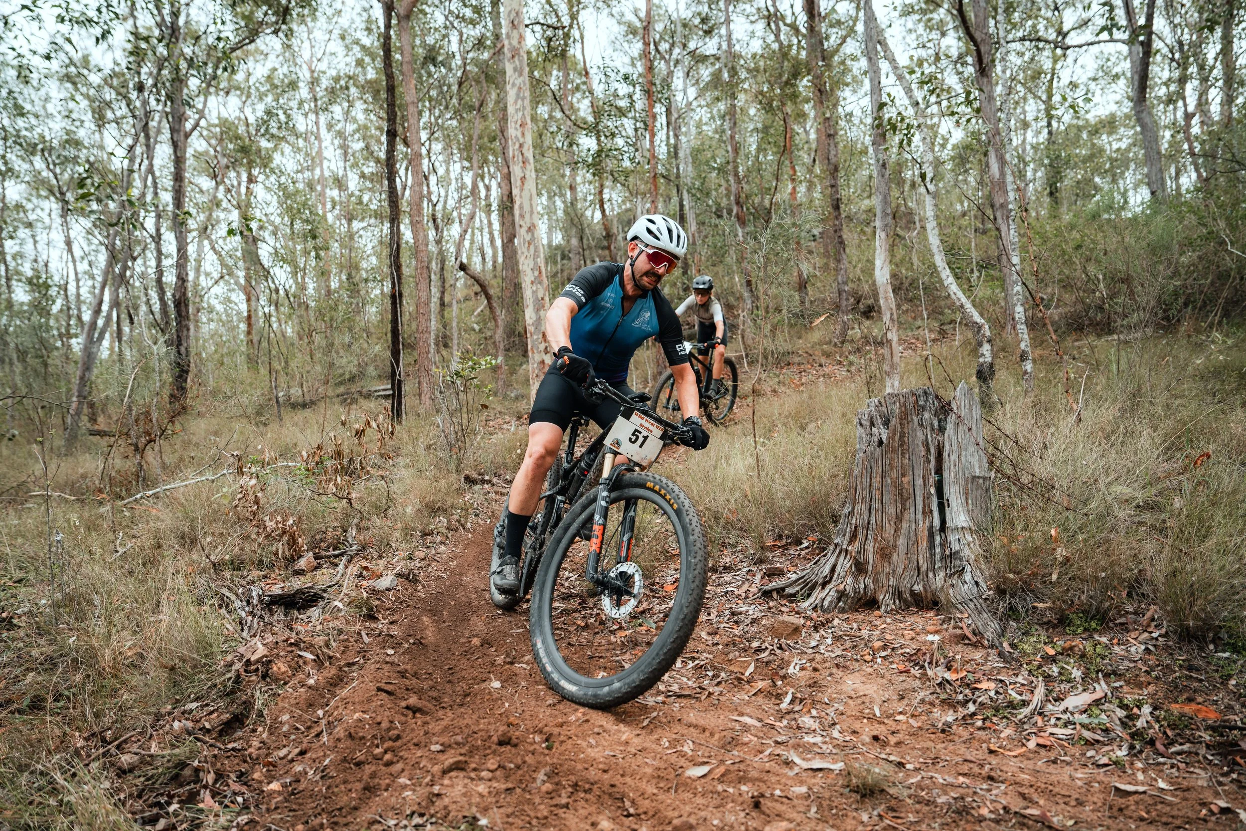 Two mountain bikers riding on a dirt trail in a forest, with trees and a tree stump nearby.