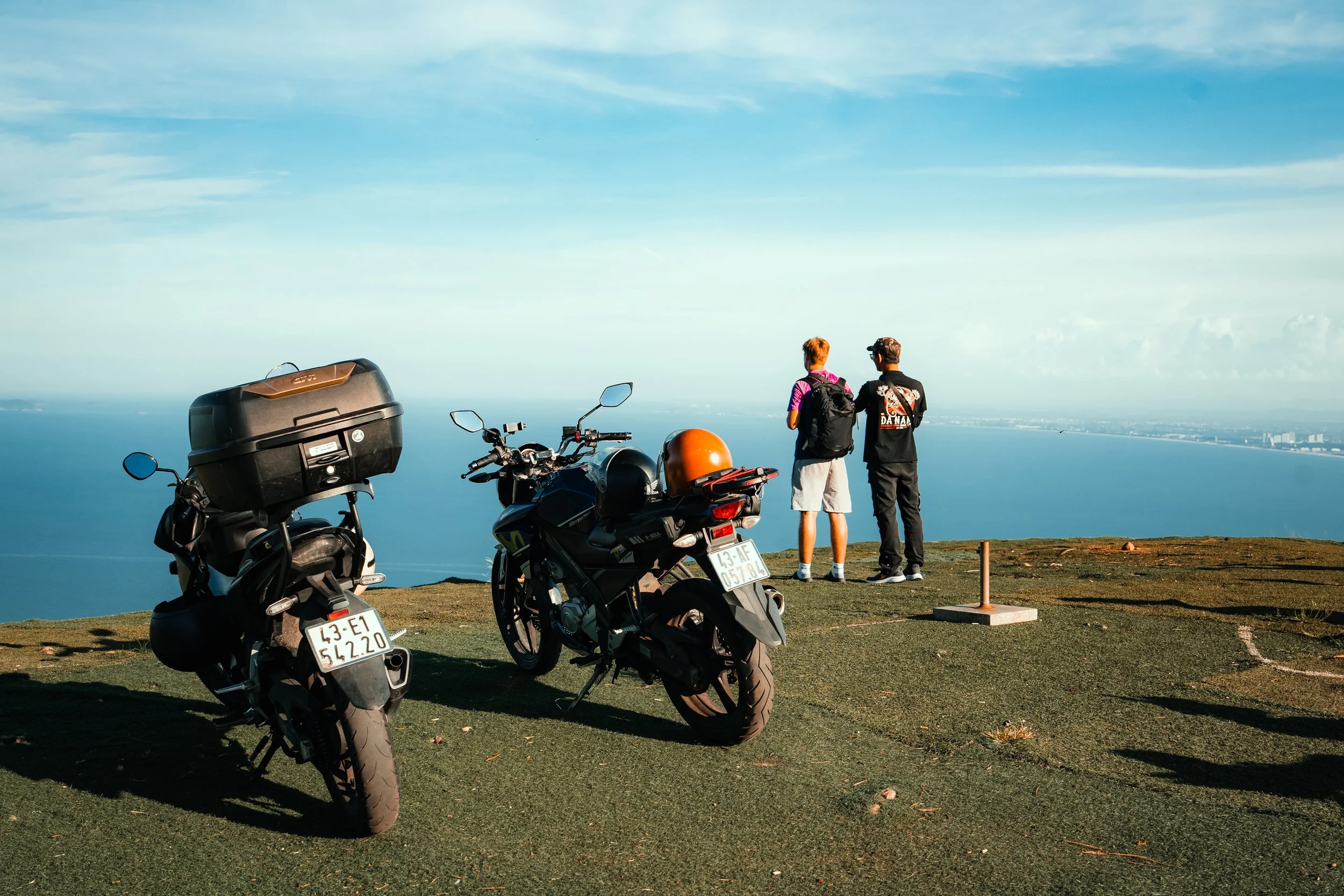 Two young men with backpacks stand on a grassy hill overlooking the ocean, with two motorcycles parked nearby.