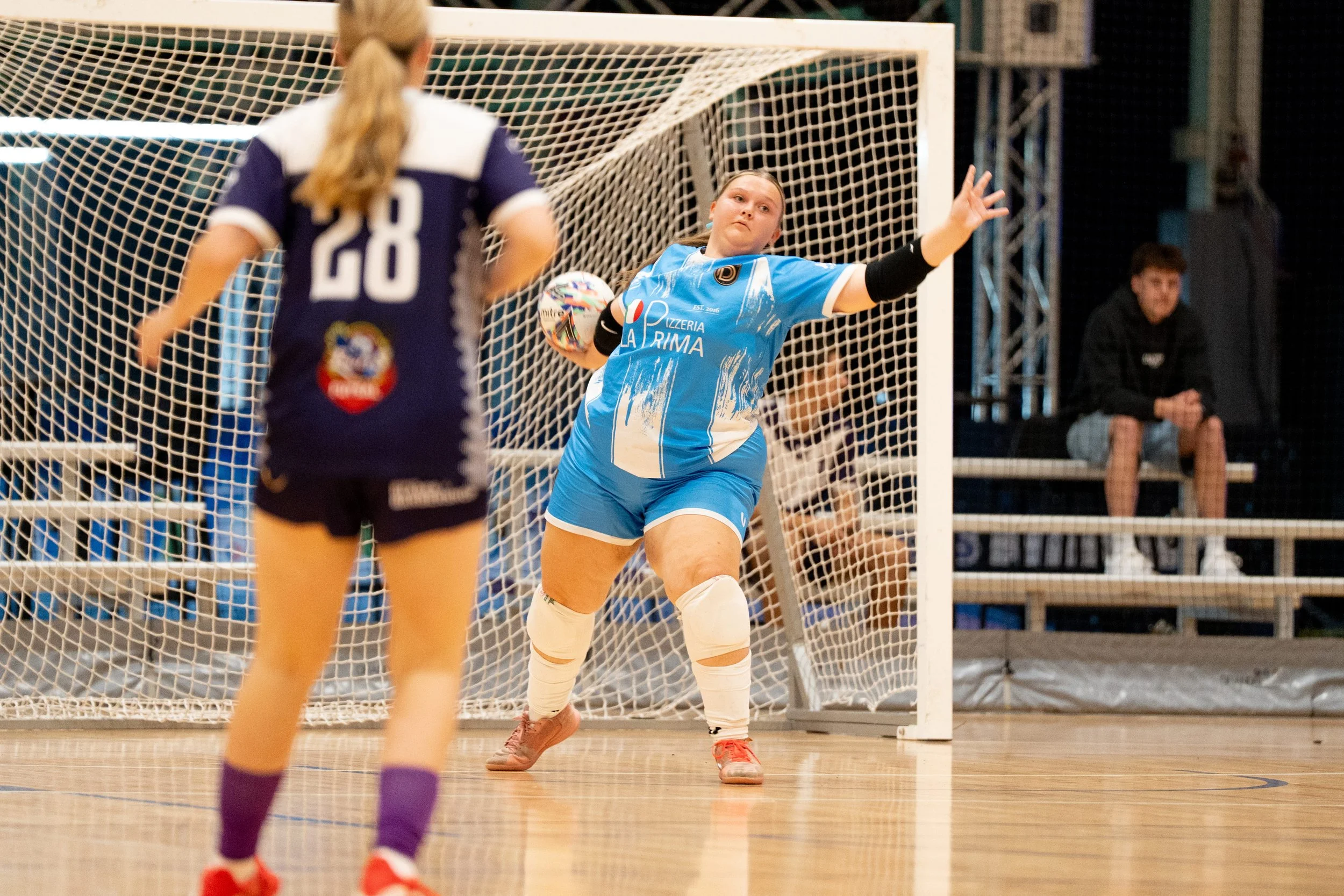 A female goalkeeper in a blue uniform with a hand band is jumping to block a shot during a indoor soccer game. A female player in a dark vein uniform with the number 28 on the back is standing in front of the goal, and a seated spectator is watching 