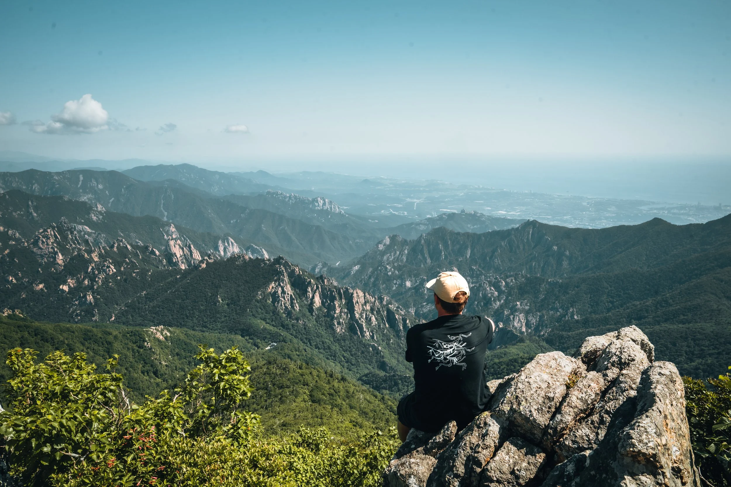 Person sitting on a rock overlooking a mountain range and distant ocean in a scenic outdoor setting.