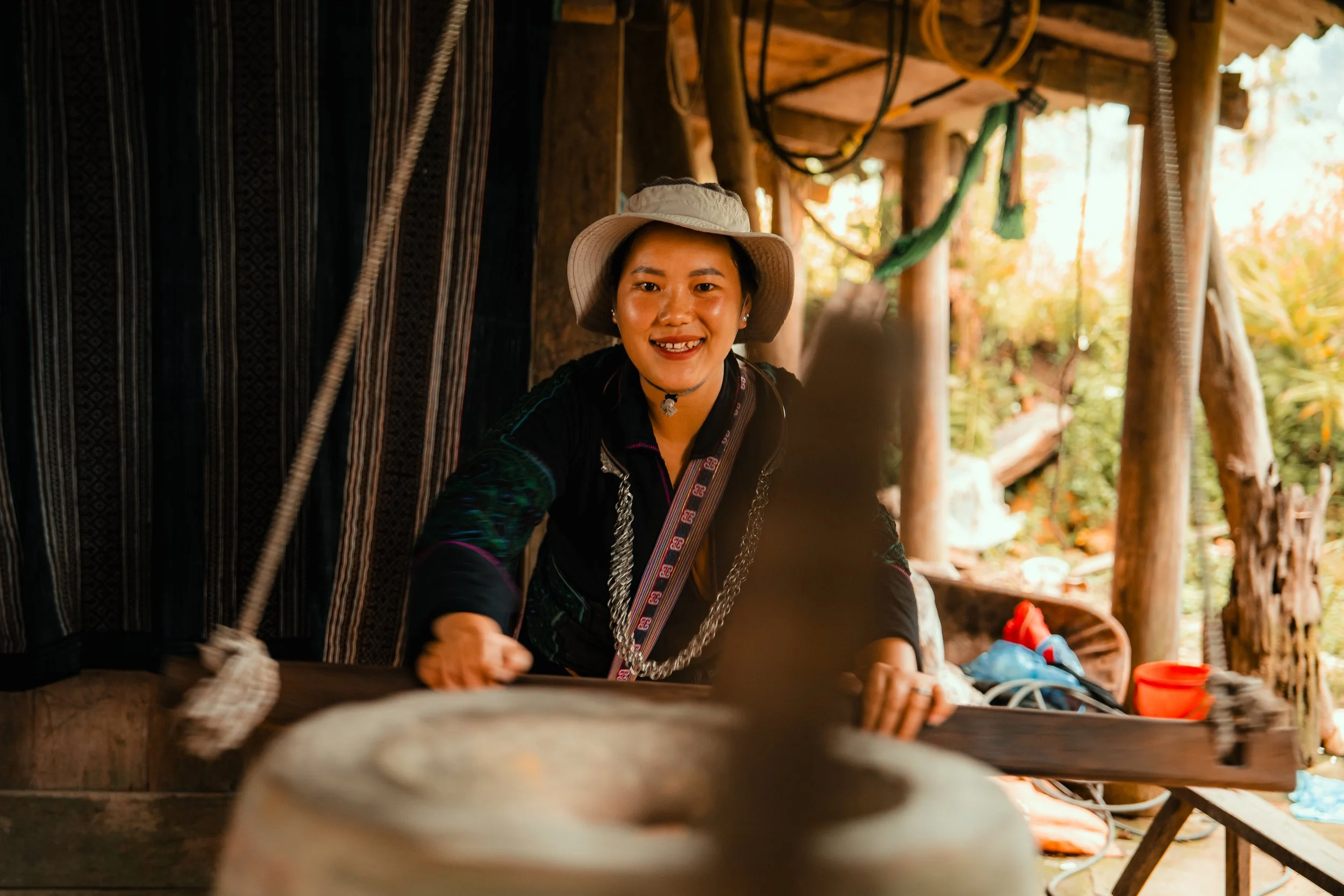 A woman in traditional attire, smiling, wearing a hat, working with a wooden tool inside a rustic wooden structure.
