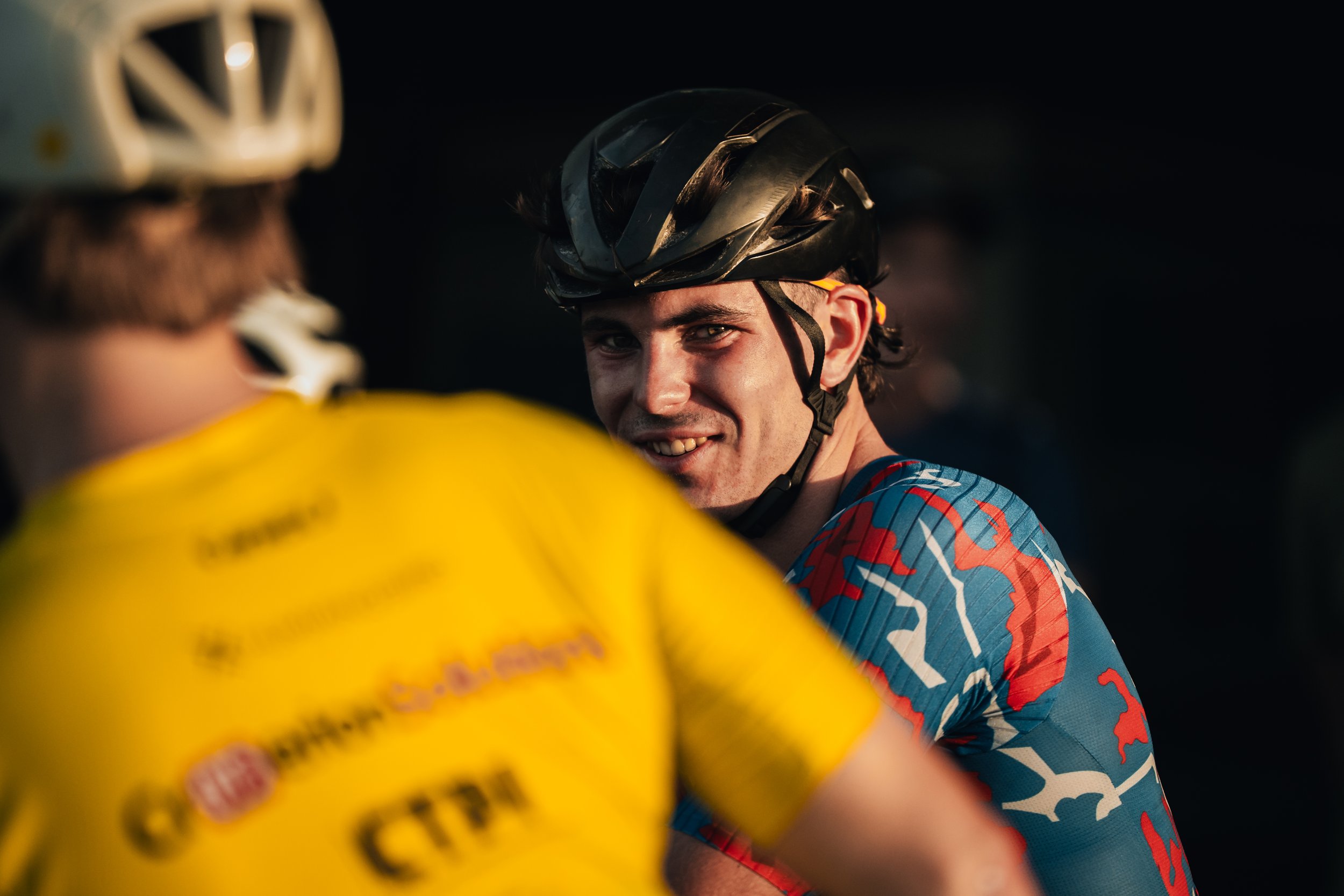 A smiling man wearing a black bicycle helmet and a colorful cycling jersey with blue, red, and white patterns, talking to another cyclist in a yellow jersey with blurred text and a white helmet, illuminated by warm sunlight against a dark background.
