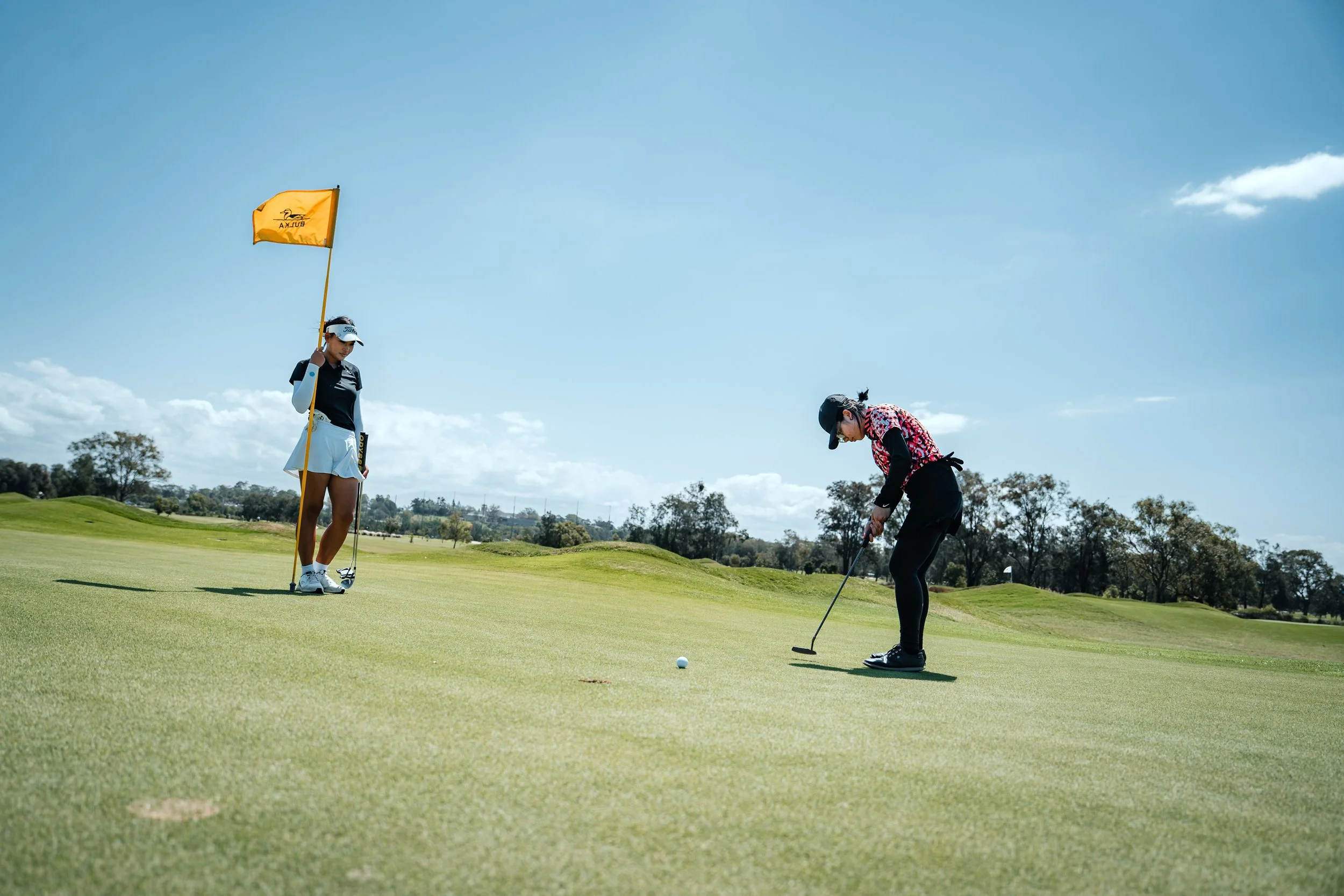 Two women playing golf on a sunny day; one is putting on the green, and the other is holding a flagstick.