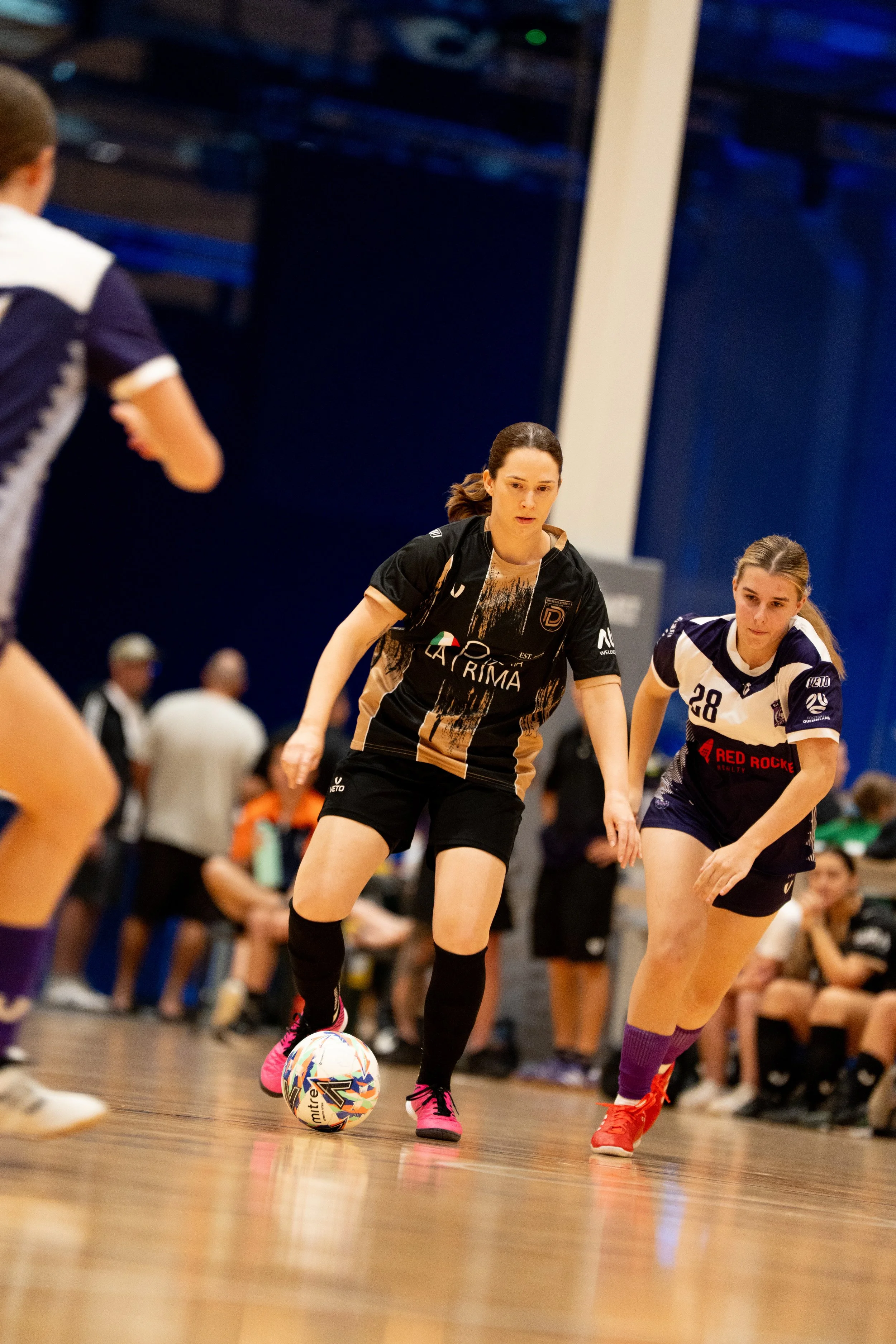 Women playing indoor soccer, one in black jersey controlling the ball while another in white and navy jersey defends, with players and spectators in the background.