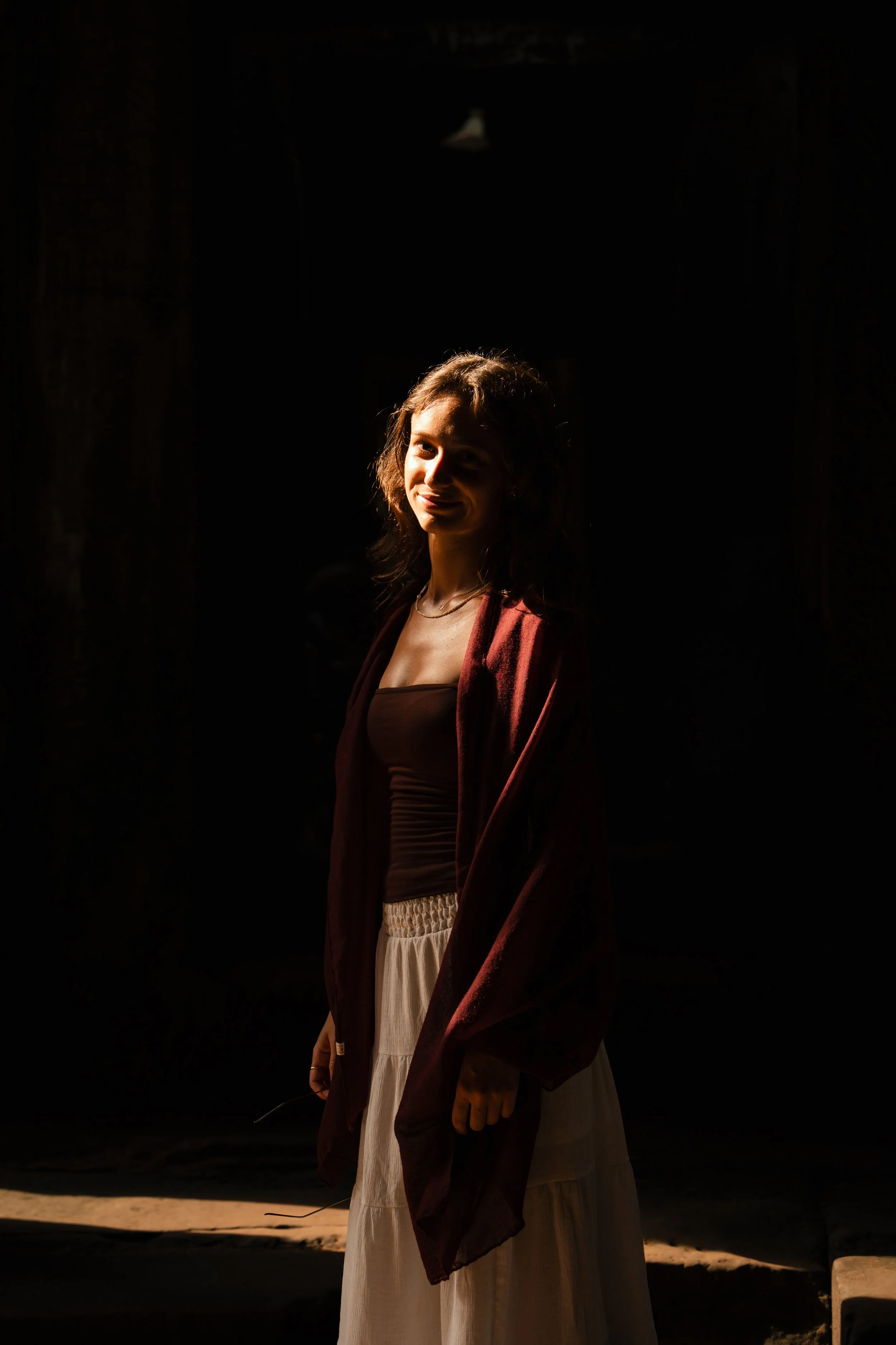 A woman standing in a dark space with sunlight illuminating her face and upper body, wearing a dark top, light-colored skirt, and a dark shawl.