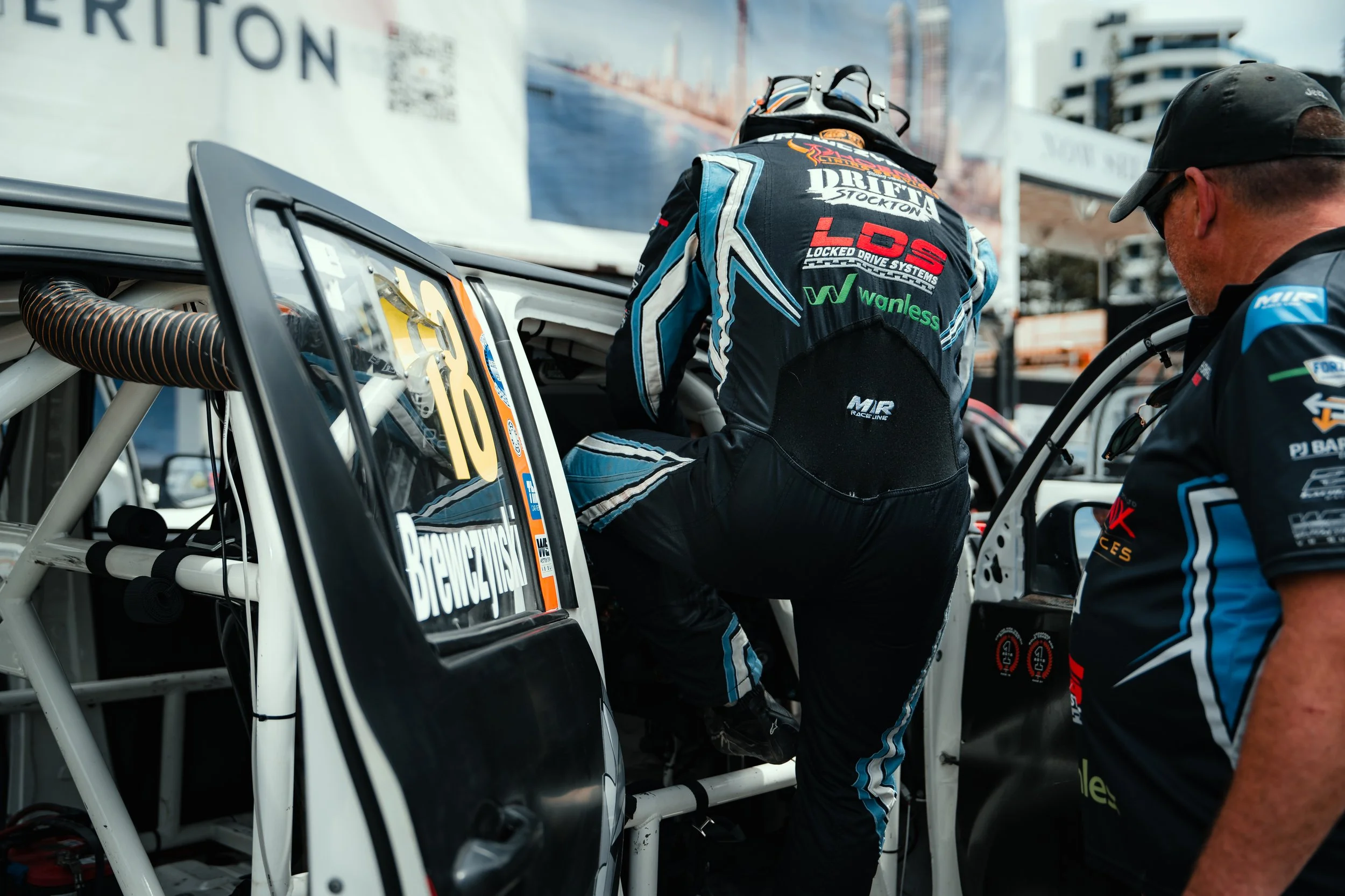 Race car driver in racing suit entering a race car, with team member standing nearby, at a racing event.