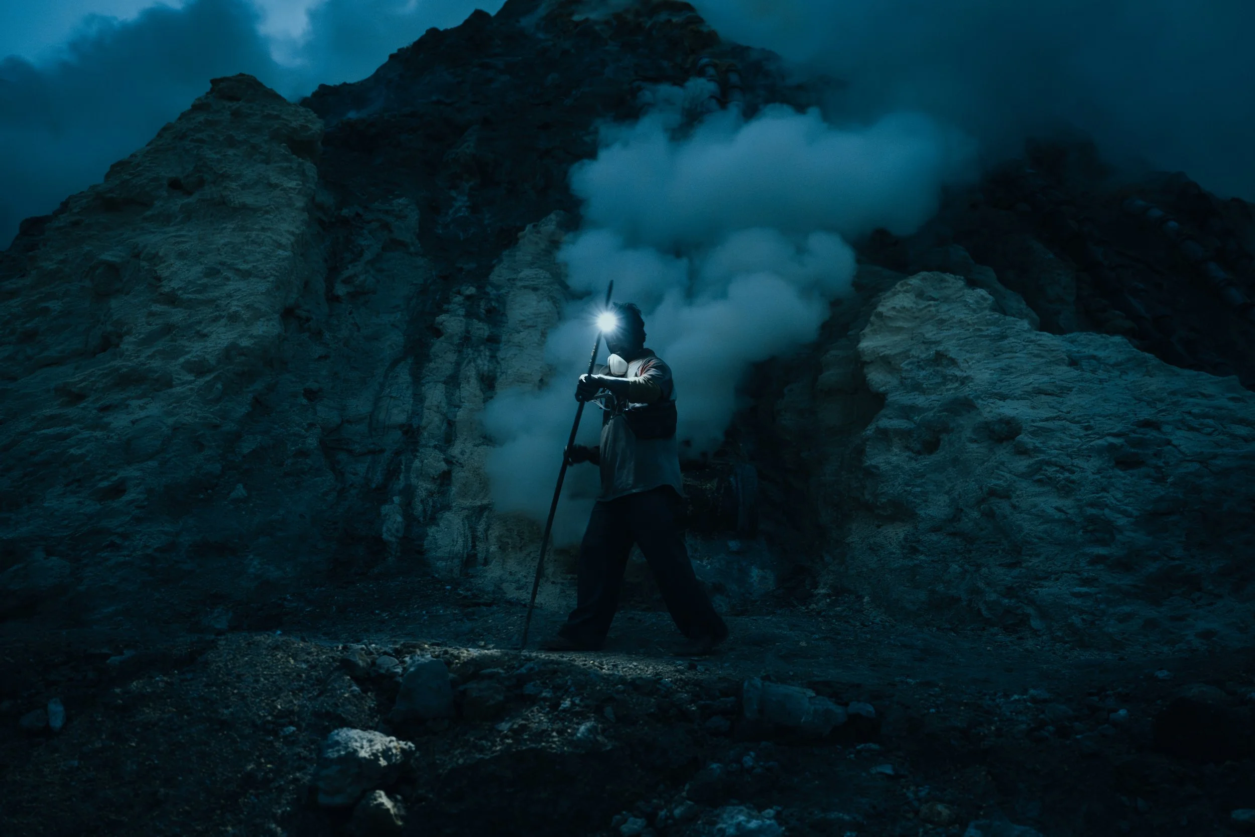 Person wearing a headlamp and face mask standing in front of a volcanic eruption with smoke and ash rising from the crater.