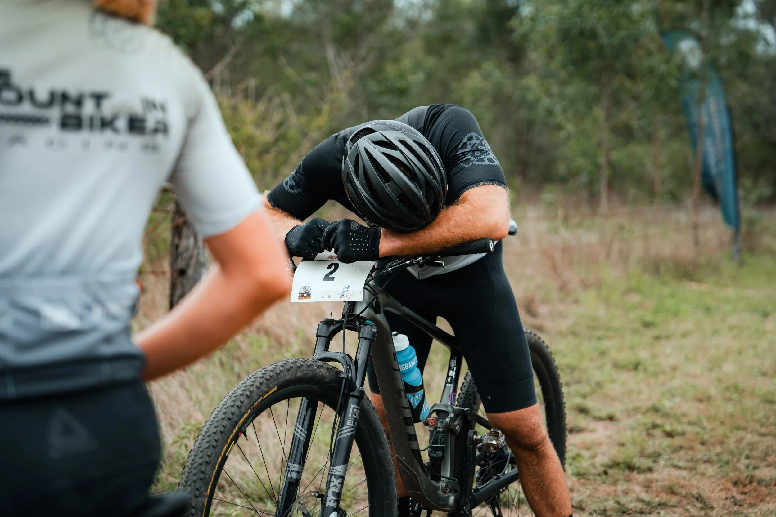 A mountain biker with a black helmet and black cycling outfit is resting with his head on his bike's handlebars during a race, with another person partially visible in the foreground and trees in the background.