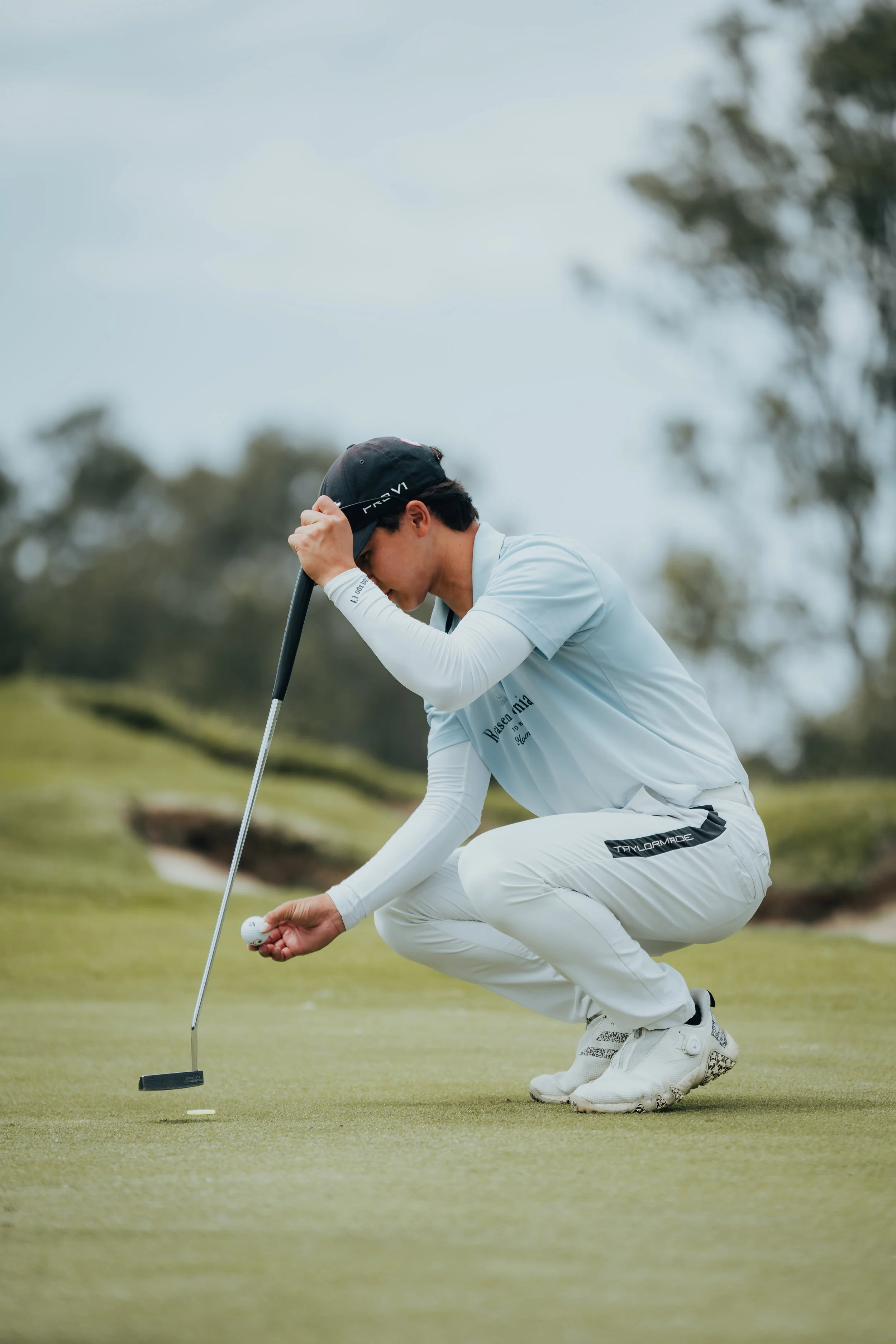 A golfer squatting on the green, lining up a putt with a golf ball and putter, on a golf course with trees and cloudy sky in background.