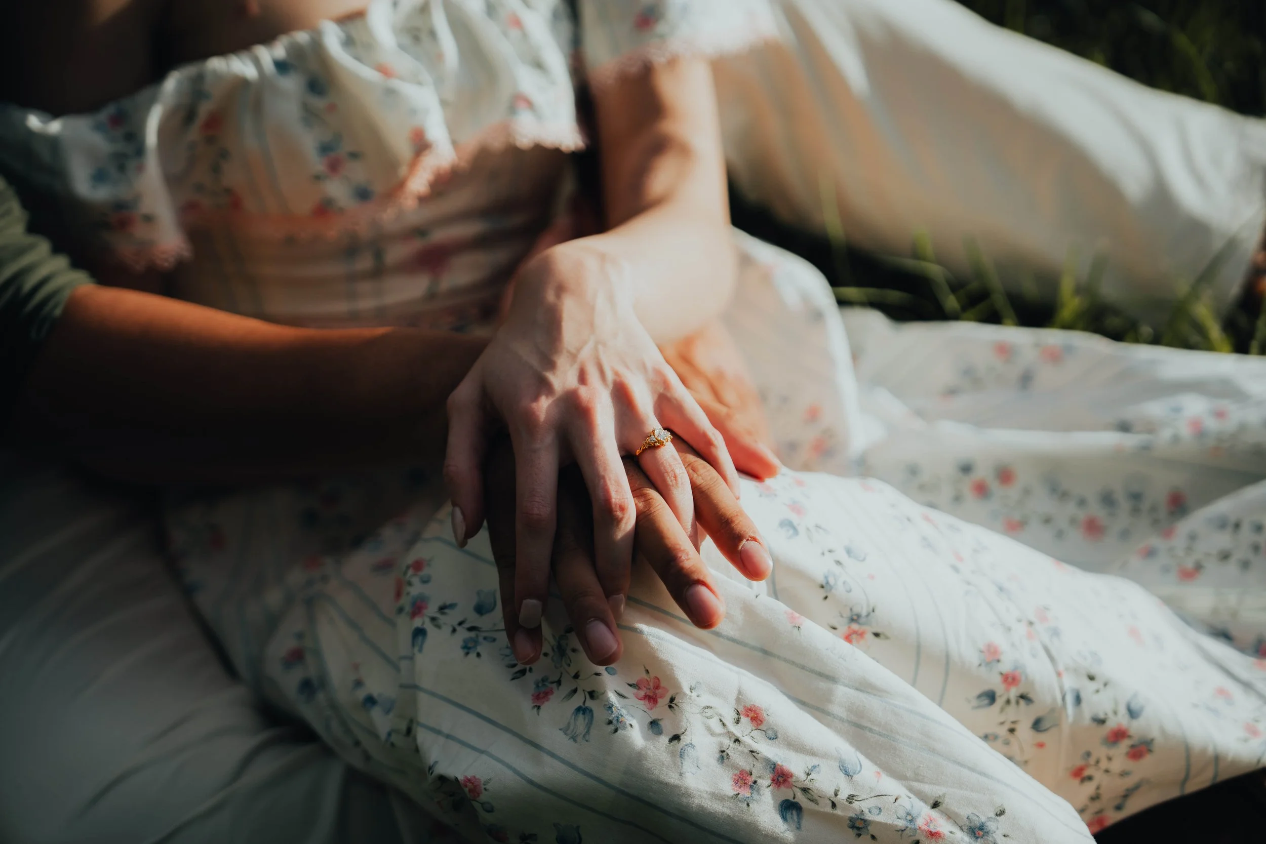 A close-up of two hands intertwined, one on top of the other, resting on a floral-patterned dress. The person is wearing a ring on their finger.