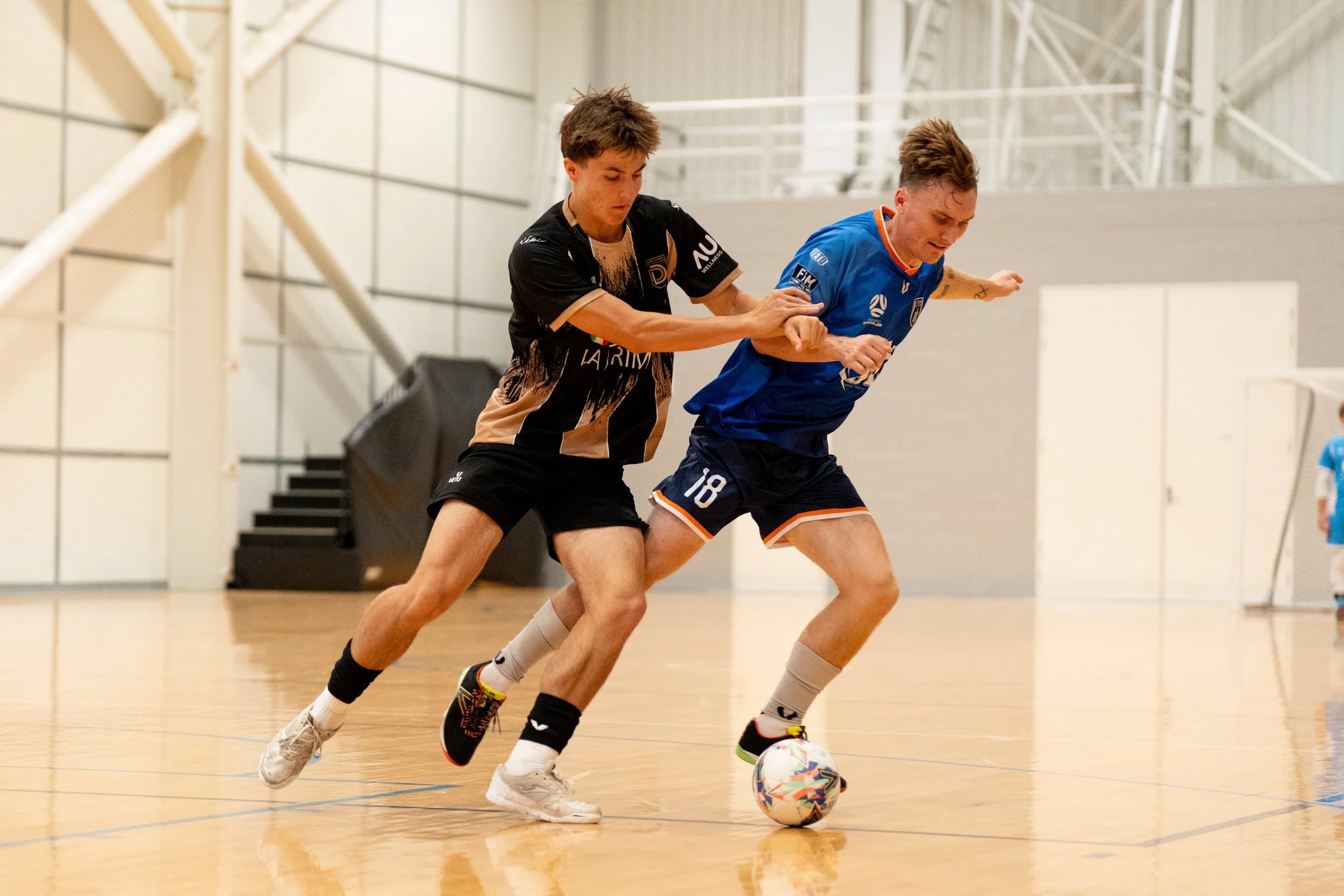 Two young men playing indoor soccer, one in black and tan jersey and the other in blue jersey, compete for the ball.