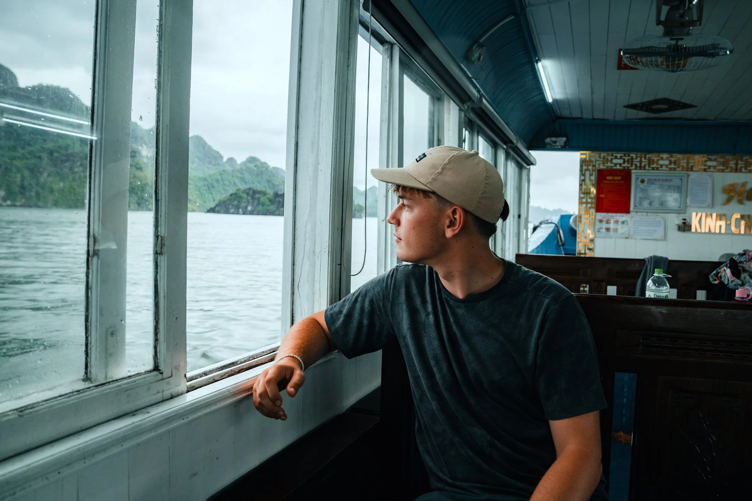 A young man in a beige cap and dark t-shirt sitting on a boat, gazing out the window at the water and green islands.