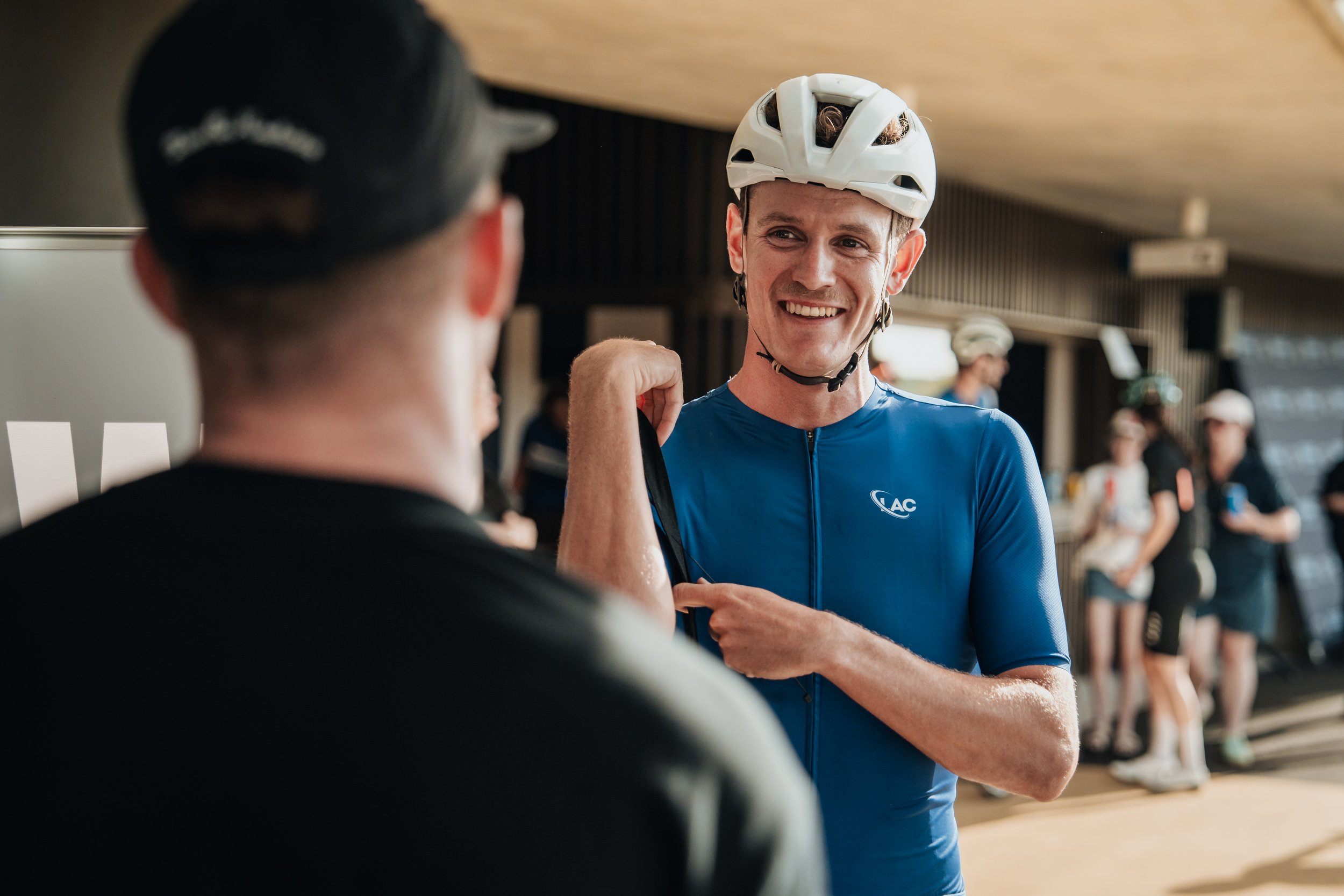 Smiling cyclist in blue jersey with helmet talking to another person at a bike event, with others in the background.