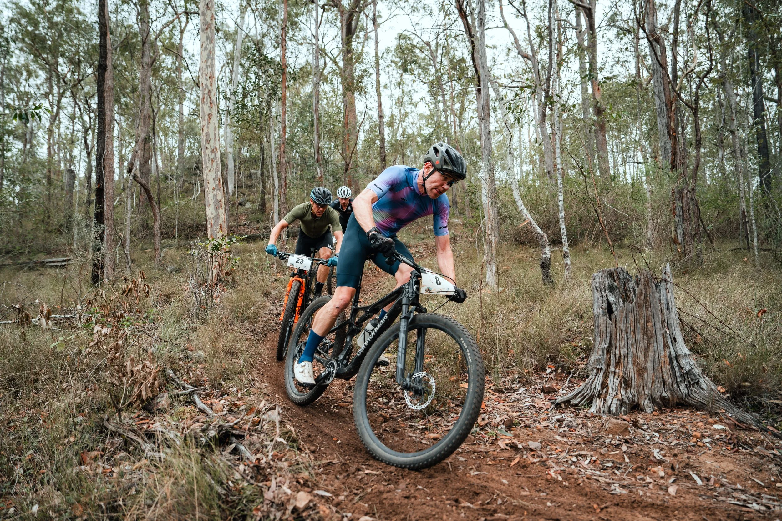 Three mountain bikers riding on a dirt trail through a wooded forest, wearing helmets and cycling gear, with one leading and the others following close behind.