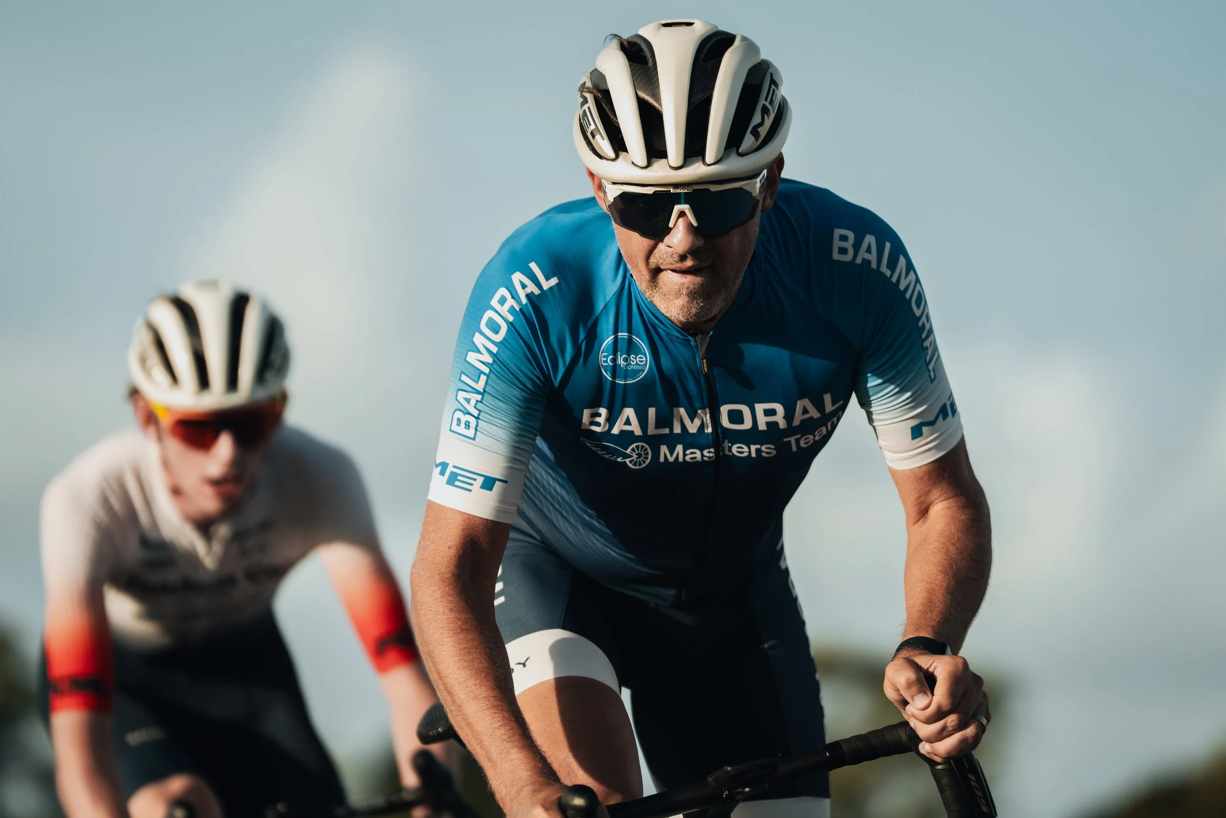 Two male cyclists wearing helmets and sunglasses racing on a sunny day, with one cyclist in a blue jersey in the foreground.