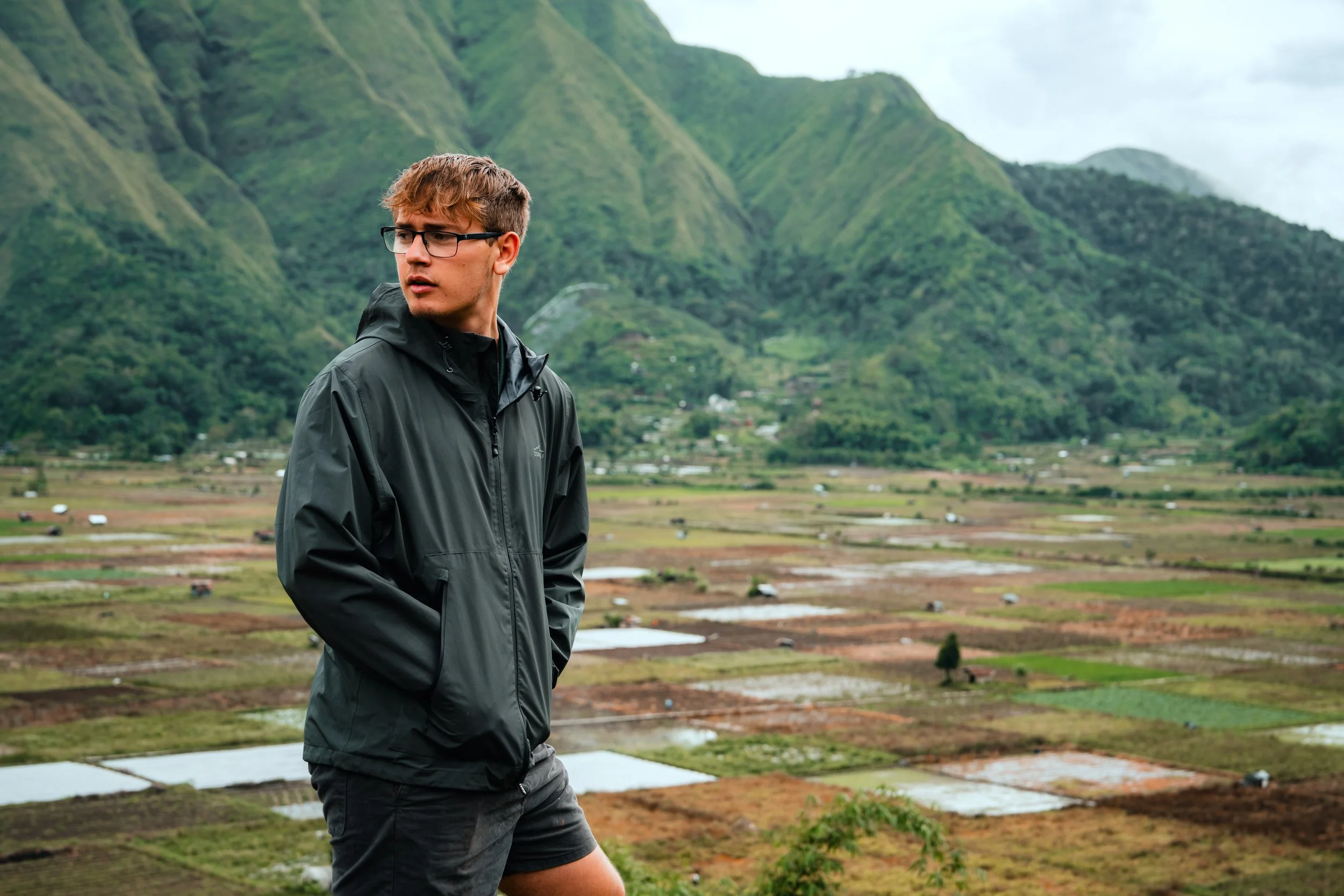 Young man in glasses and a black windbreaker stands outdoors with a mountainous landscape and patchwork fields in the background.
