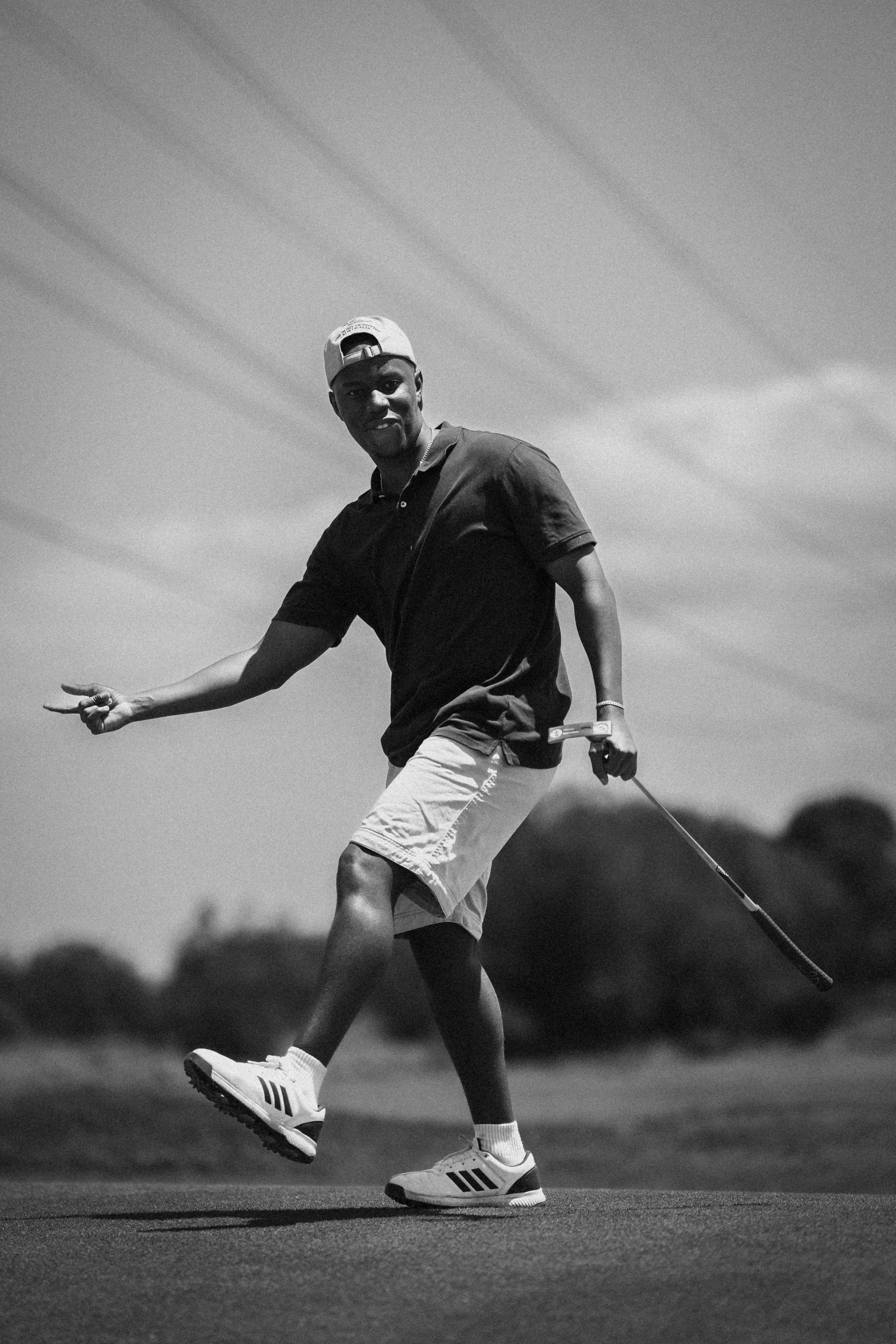 A young man in shorts, t-shirt, and athletic shoes walking on a golf course while holding a golf club, smiling and making a peace sign with his left hand, under an overcast sky with faint clouds.